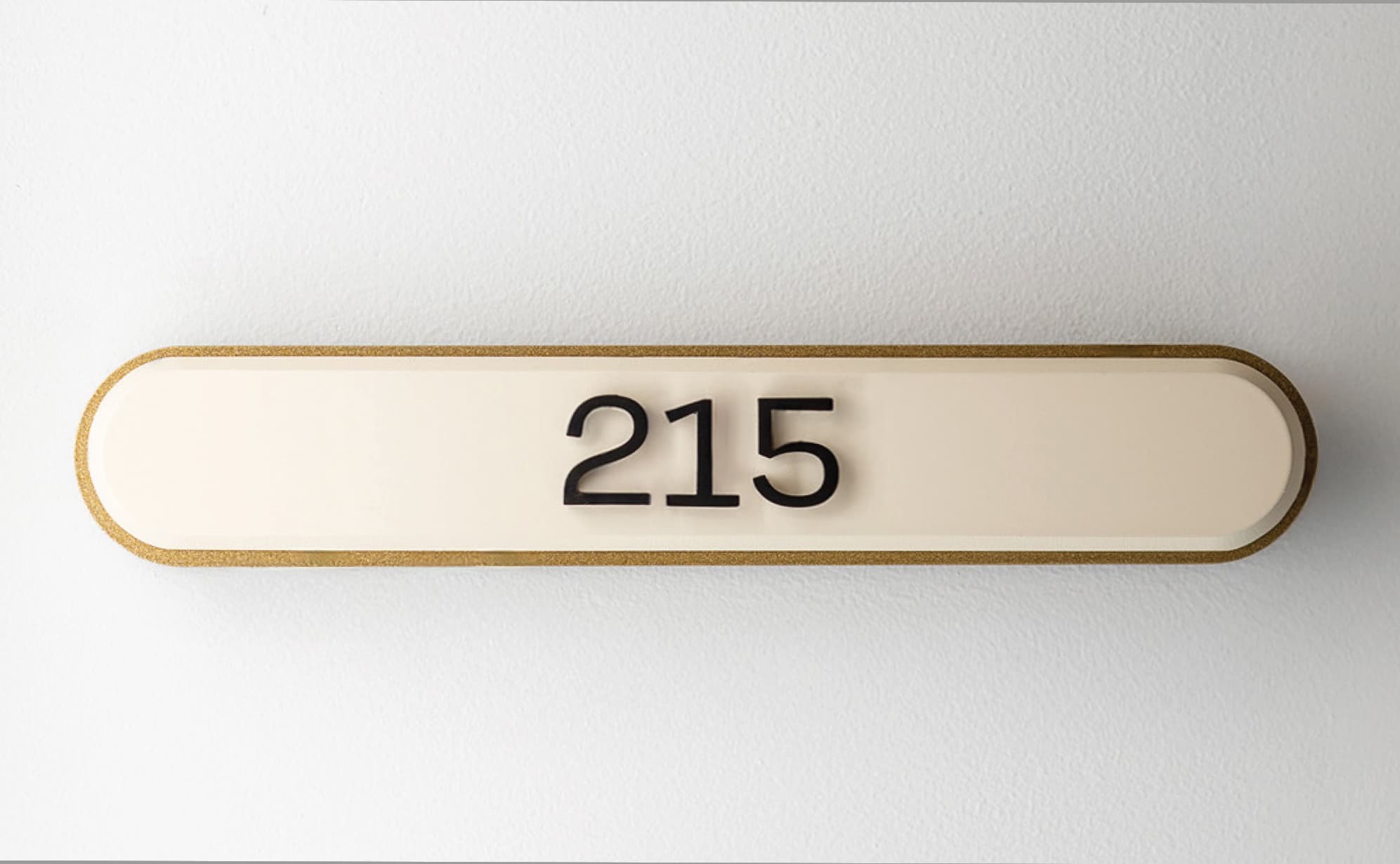 RSM Design interior unit identification signage for The Charles at The West Quarter in Salt Lake City, featuring a close-up of a cream pill-shaped room number plaque with dimensional black numerals and a gold glitter edge detail mounted on a white wall, showcasing how specialty graphics, environmental graphic design, and master sign program design extend visual identity and branded environment cohesion to every interior touchpoint within mixed-use hospitality and residential community markets.