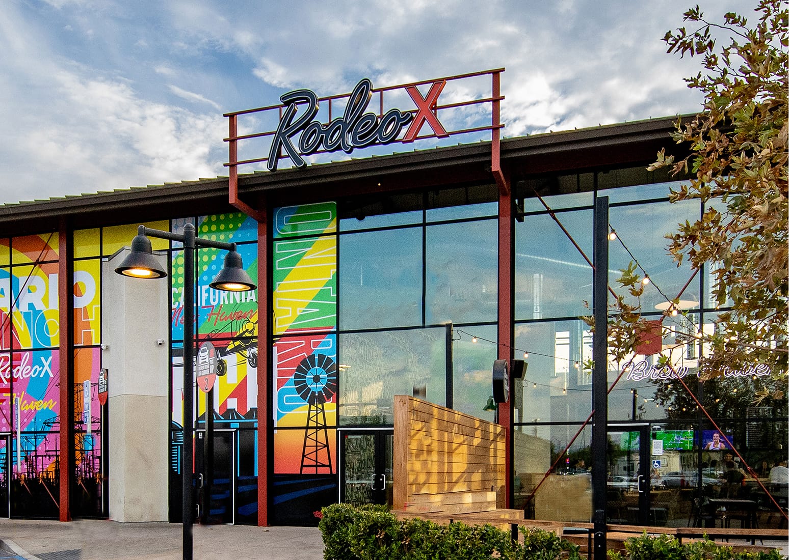 Glass-walled 'Rodeo X' building with a rooftop script sign, colorful window graphics depicting Ontario Ranch imagery like a windmill and power lines, string lights, and a wood accent wall at the entrance.