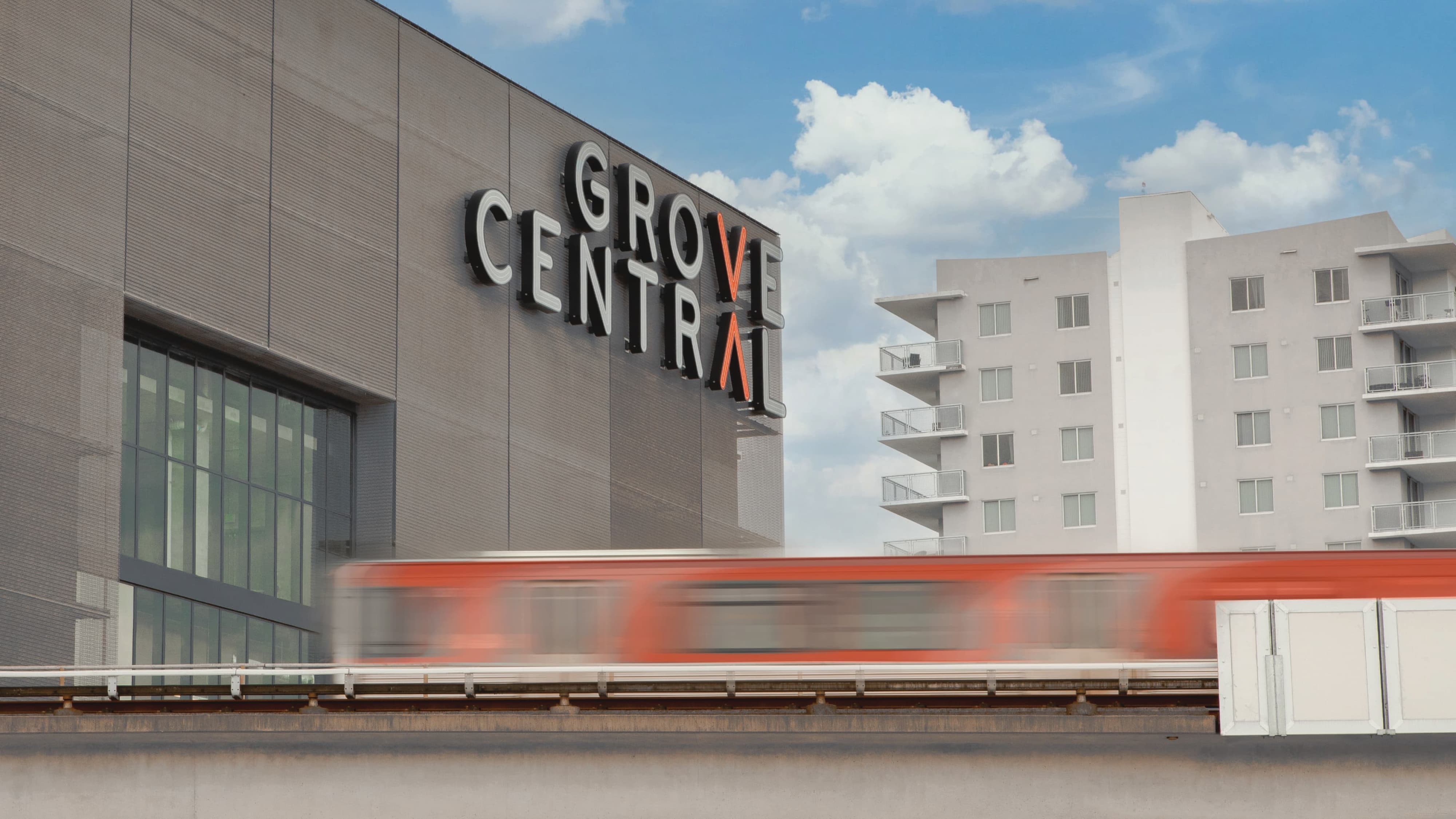 Grove Central master sign mounted on a building facade above an elevated Metrorail track, with a red and silver train passing in motion blur and residential towers visible in the background under a partly cloudy sky.