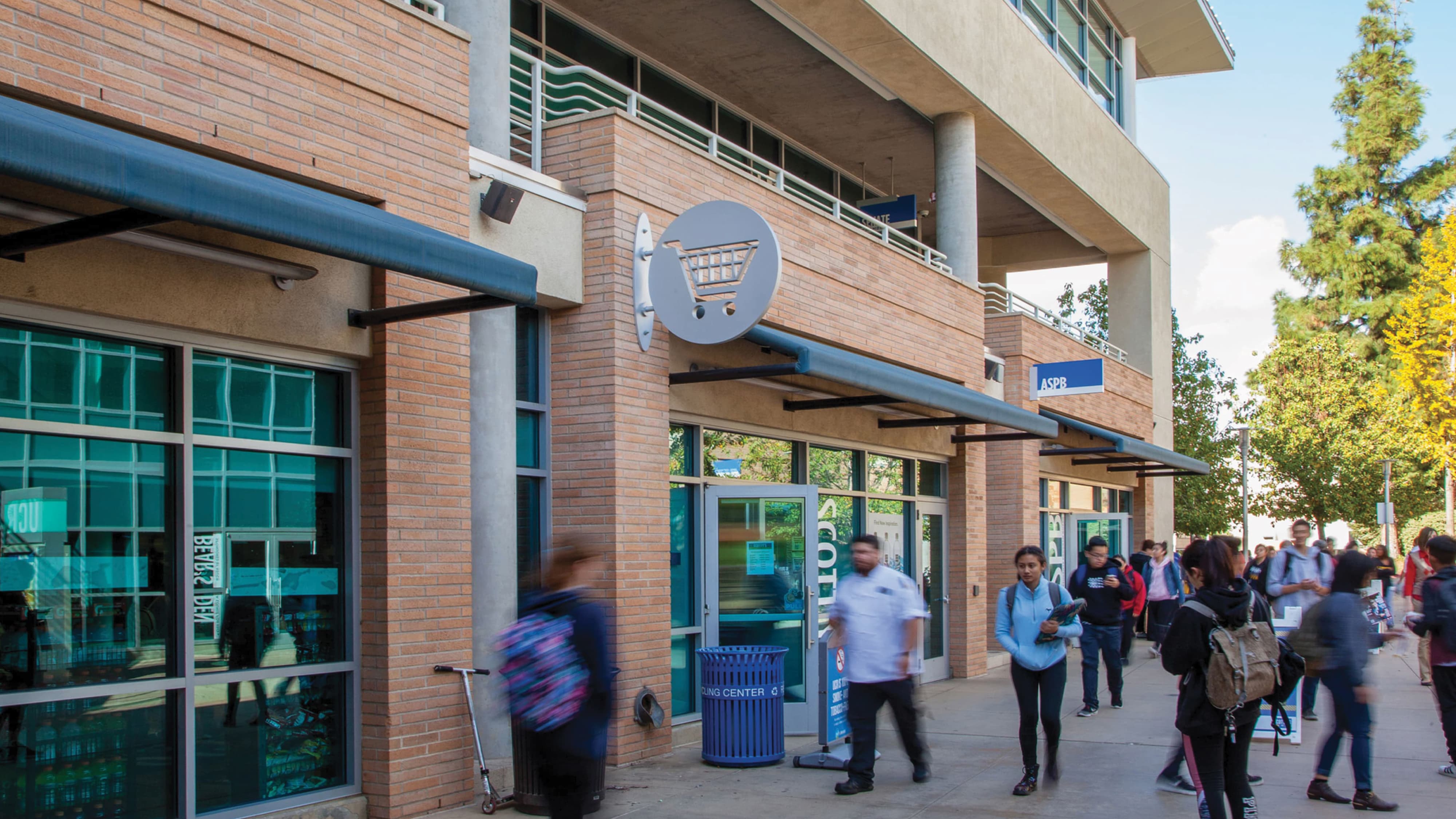Busy campus sidewalk beneath a brick building with a shopping cart icon sign and 'ASPB' storefront signage, students walking past a scooter and recycling bin under blue awnings.