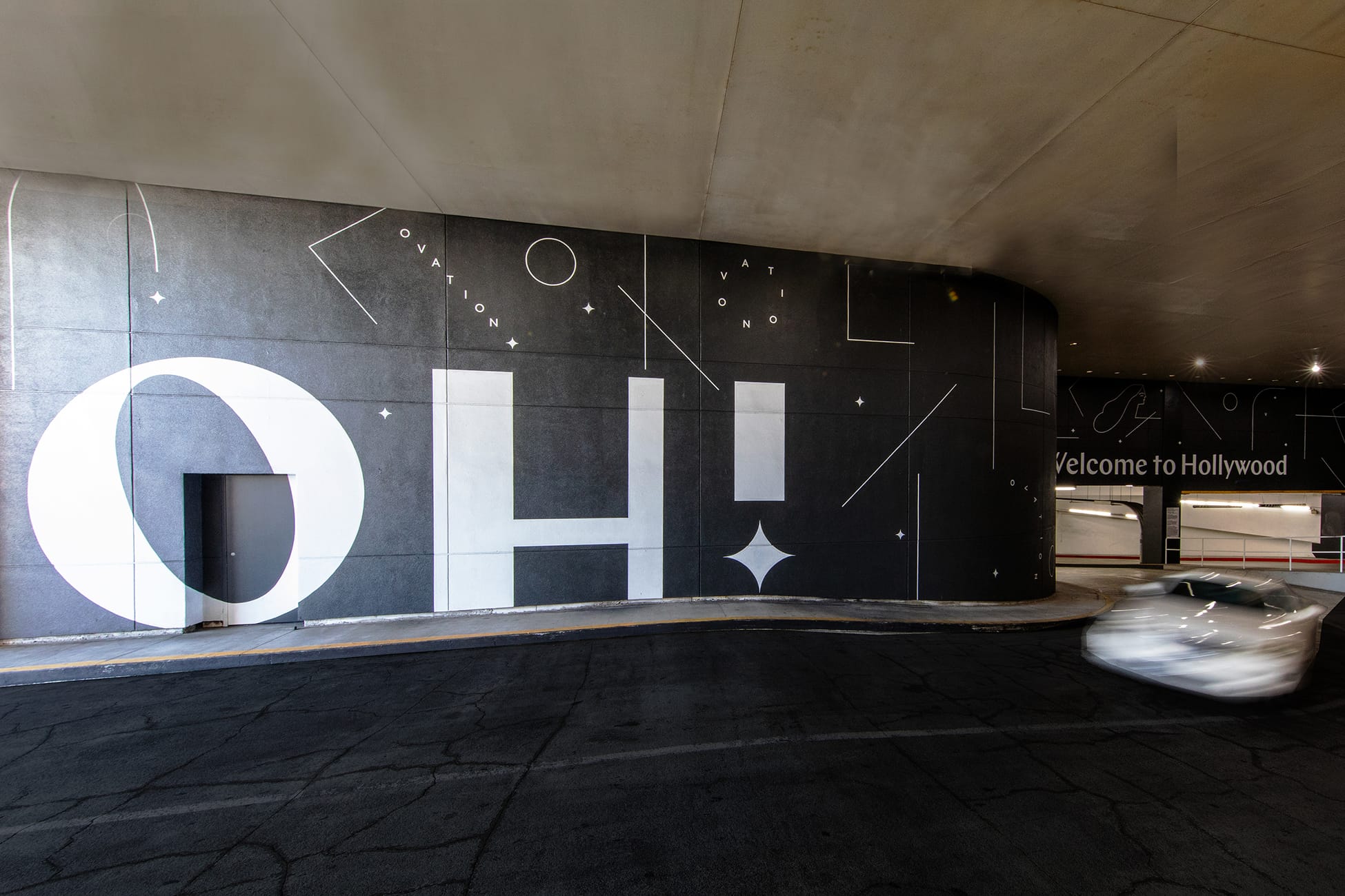 Wide-angle interior view of the Ovation Hollywood parking garage entry level featuring a sweeping curved dark charcoal wall with a dramatic large-scale black and white "OH!" typographic brand mural incorporating geometric line art, star and diamond motifs, scattered "OVATION" lettering fragments, and a "Welcome to Hollywood" greeting inscription on the curved ramp wall, with a motion-blurred vehicle navigating the entry ramp beneath an exposed concrete ceiling, creating an immersive branded arrival experience at Hollywood Boulevard Los Angeles