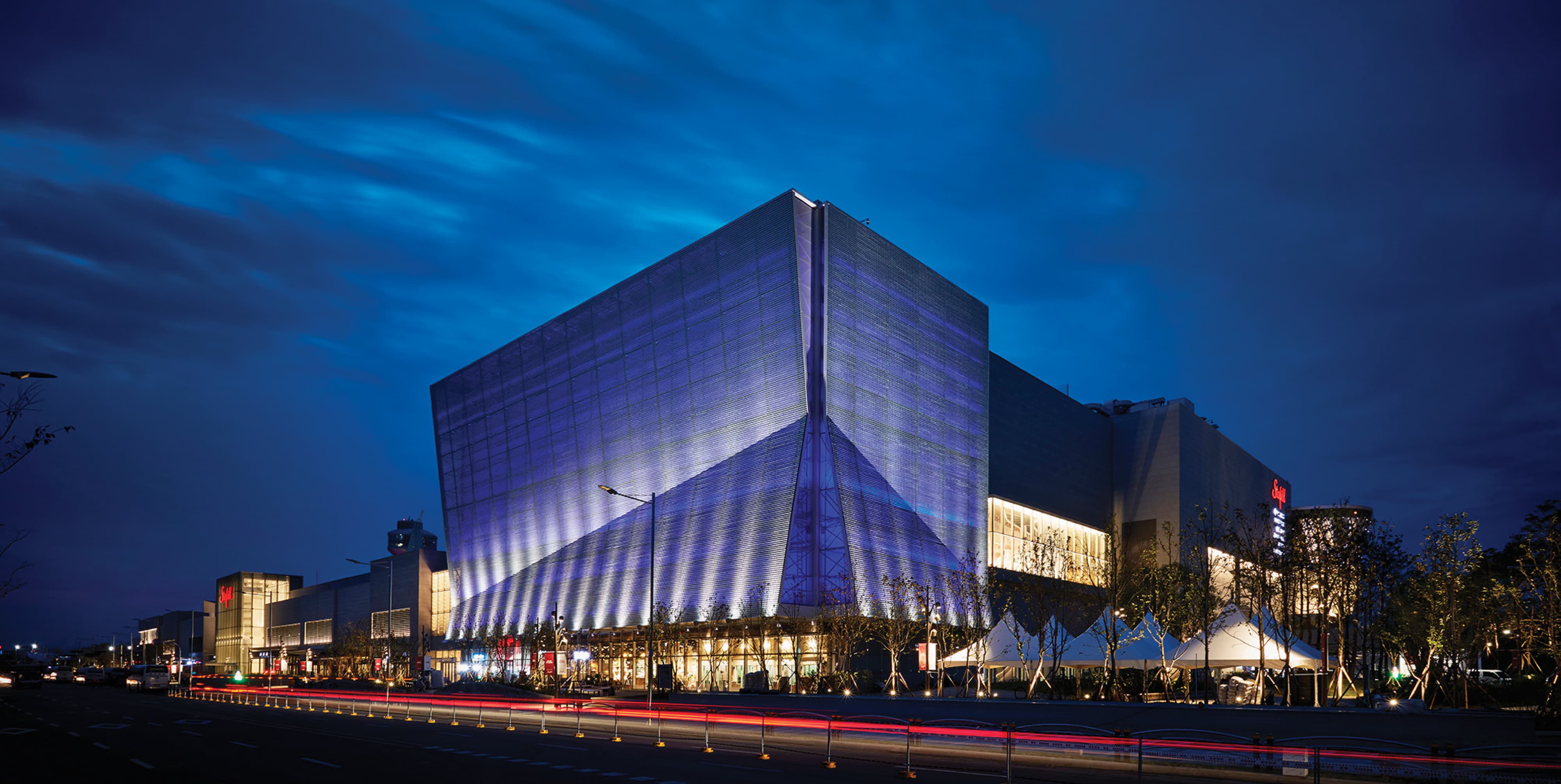 Nighttime exterior of a large geometric mall building with an angular perforated metal facade illuminated in blue, storefronts and canopy tents lit at ground level, and light trails from passing traffic on the street below.