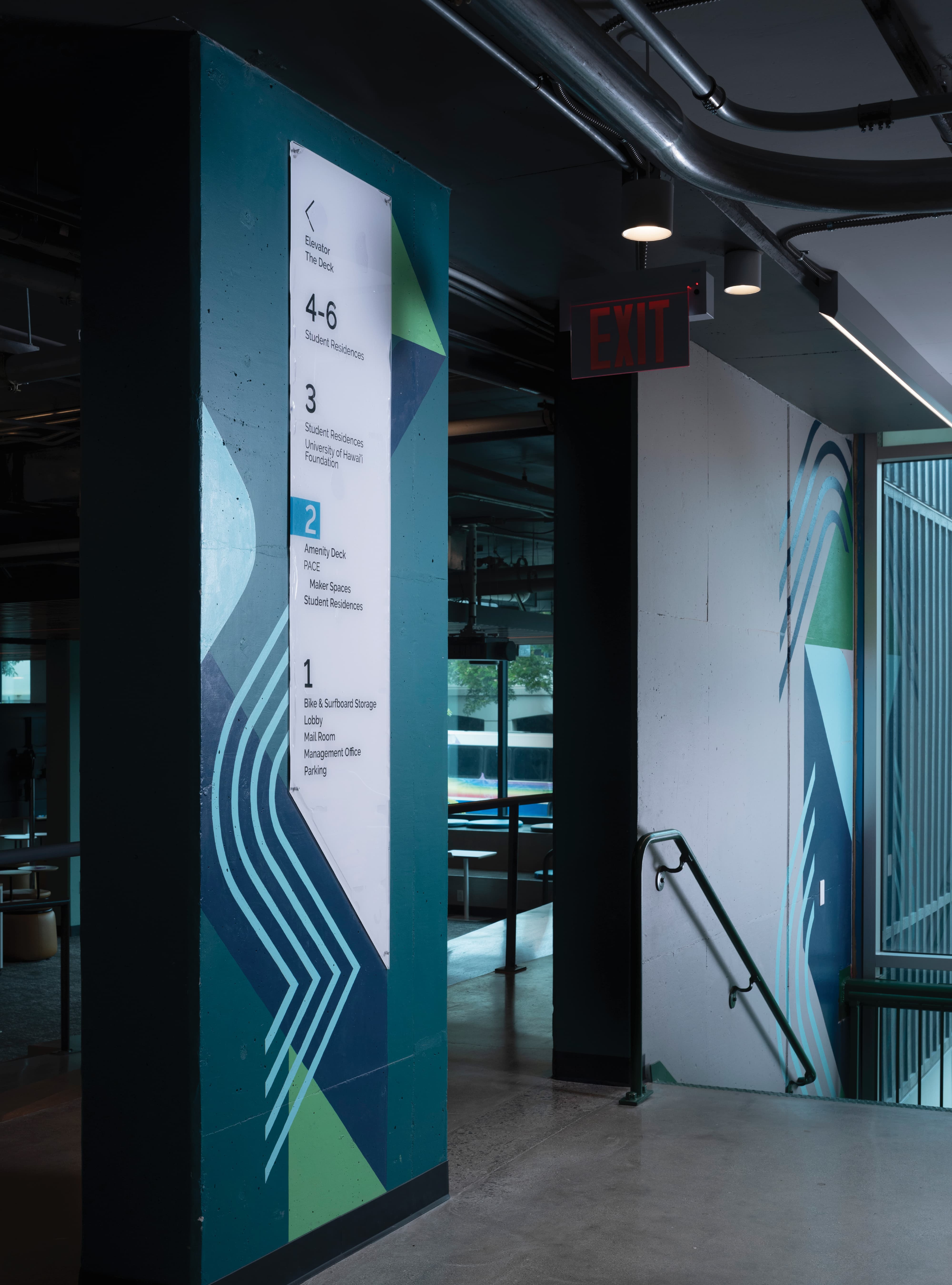 Building directory sign listing floors 1 through 6 (Bike & Surfboard Storage, Lobby, Amenity Deck, Student Residences, University of Hawaiʻi Foundation) mounted on a teal and green geometric-painted column, beside a stairwell with a matching wave-pattern mural and a lit red 'Exit' sign.