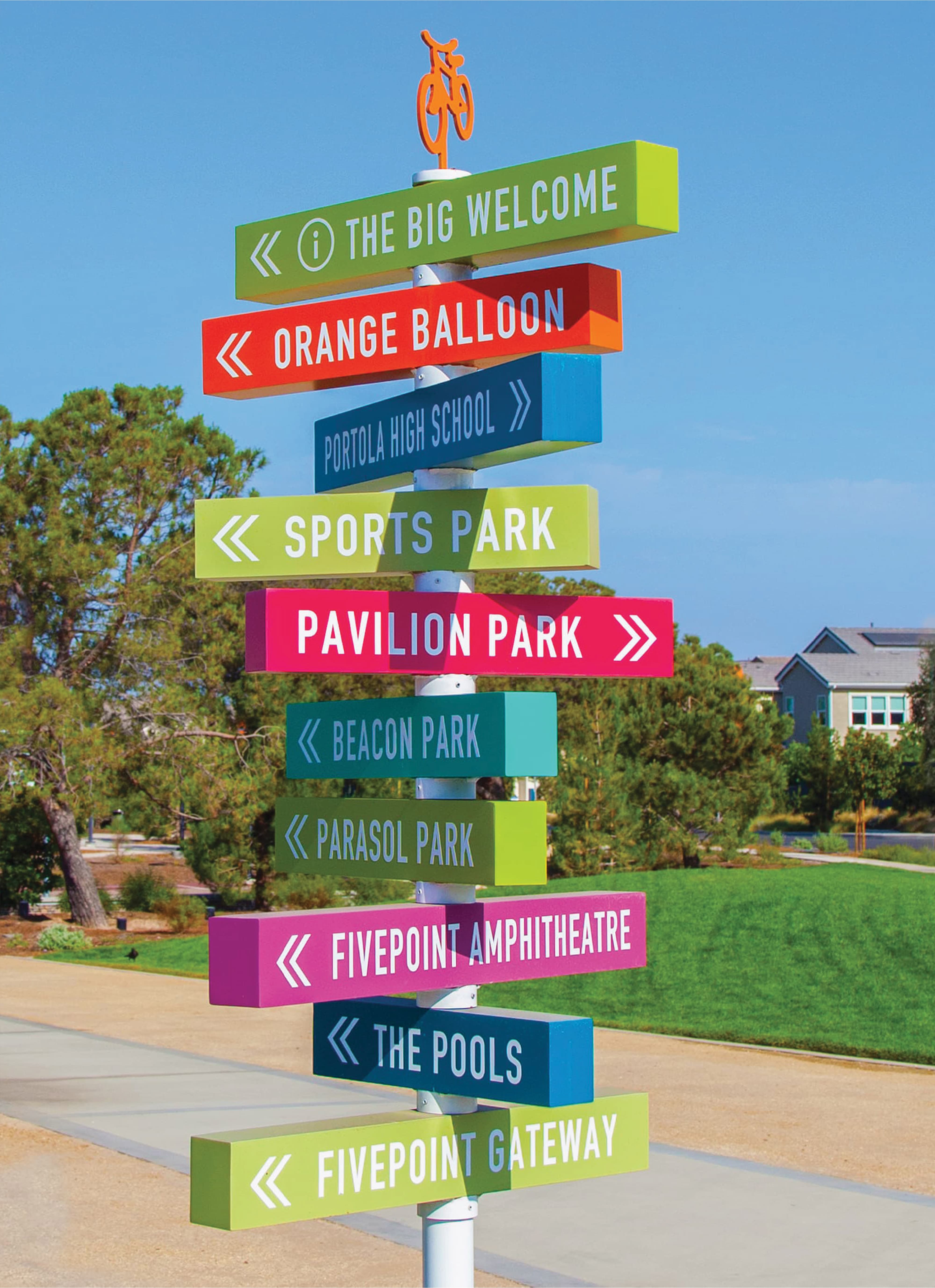 Full view of a multicolored directional wayfinding sign post topped with an orange bicycle icon, guiding residents to destinations including Pavilion Park, Beacon Park, and FivePoint Amphitheatre in a mixed-use residential community, featuring placemaking and signage design by RSM Design.