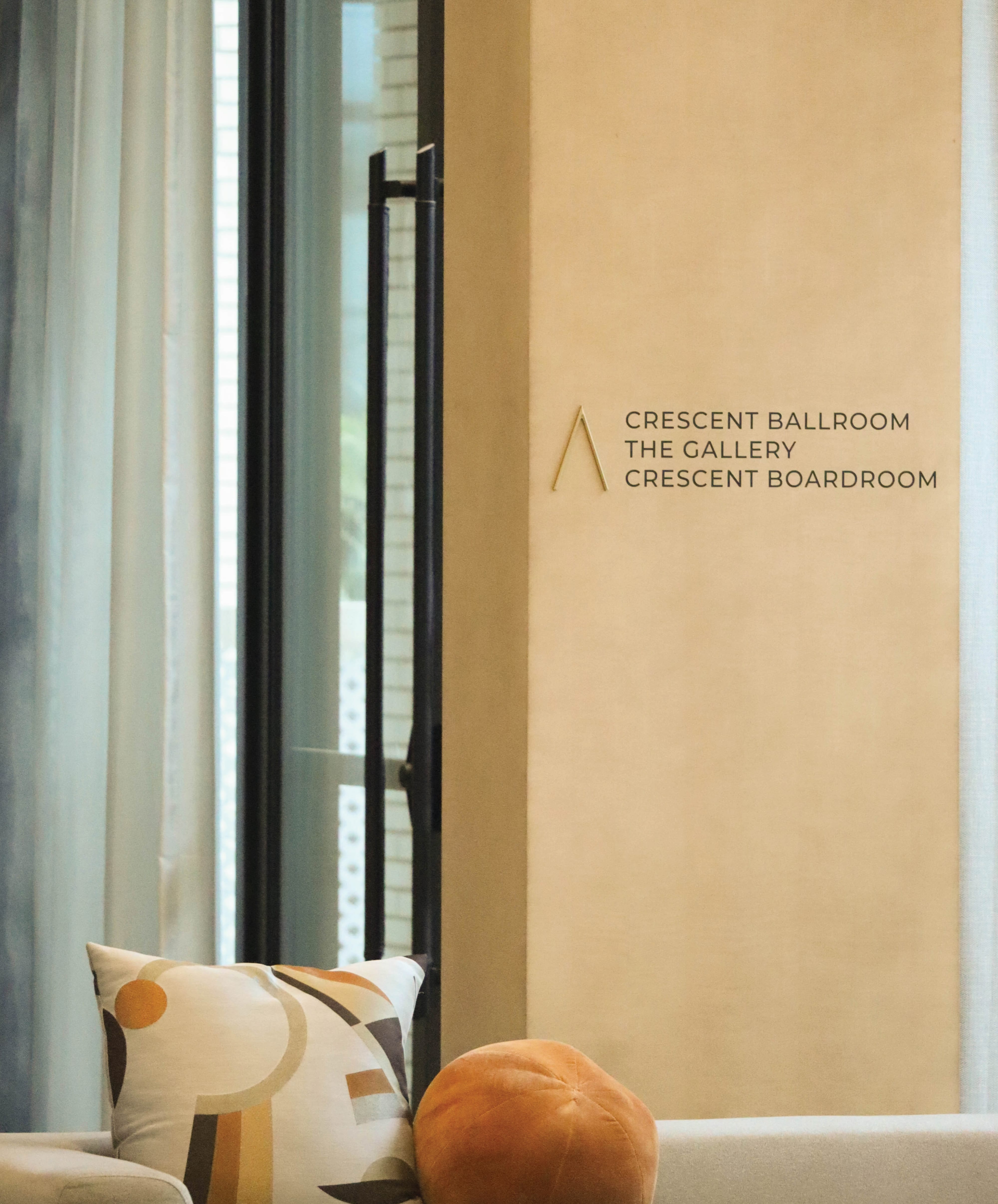 Crescent Hotel event venue directional signage pointing to Crescent Ballroom, The Gallery, and Crescent Boardroom meeting spaces, with modern geometric throw pillows and floor-to-ceiling windows in the foreground