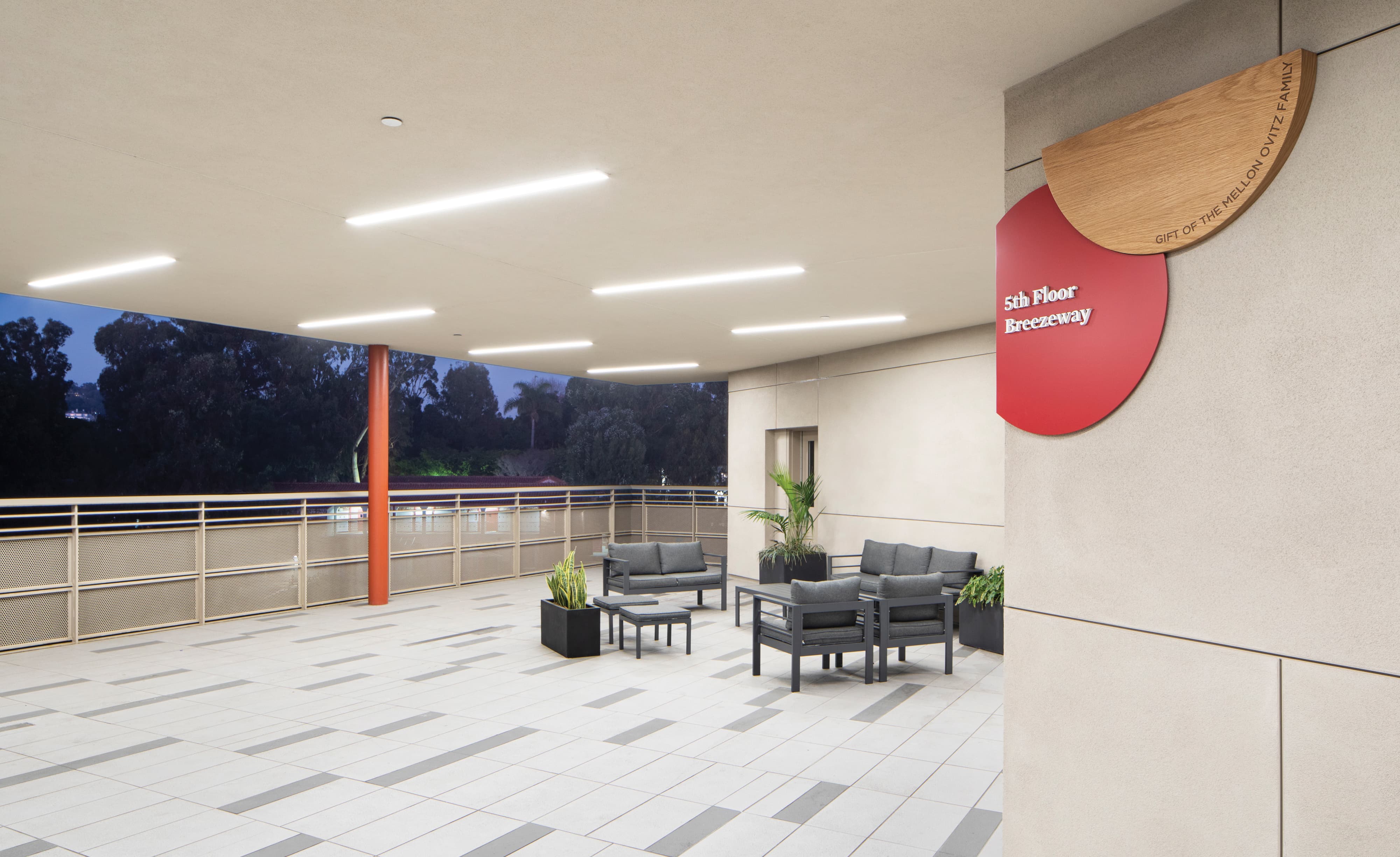 An open-air 5th Floor Breezeway lounge on an educational campus at dusk, featuring a red circular space identification sign with dimensional white lettering overlapping a natural oak wood half-circle engraved "Gift of the Mellovitz Family," mounted on a concrete column above a seating area with dark outdoor furniture, potted plants, and a tree-lined evening skyline beyond — demonstrating RSM Design's donor recognition and specialty graphics and placemaking services that create meaningful, human-centered moments within the education market