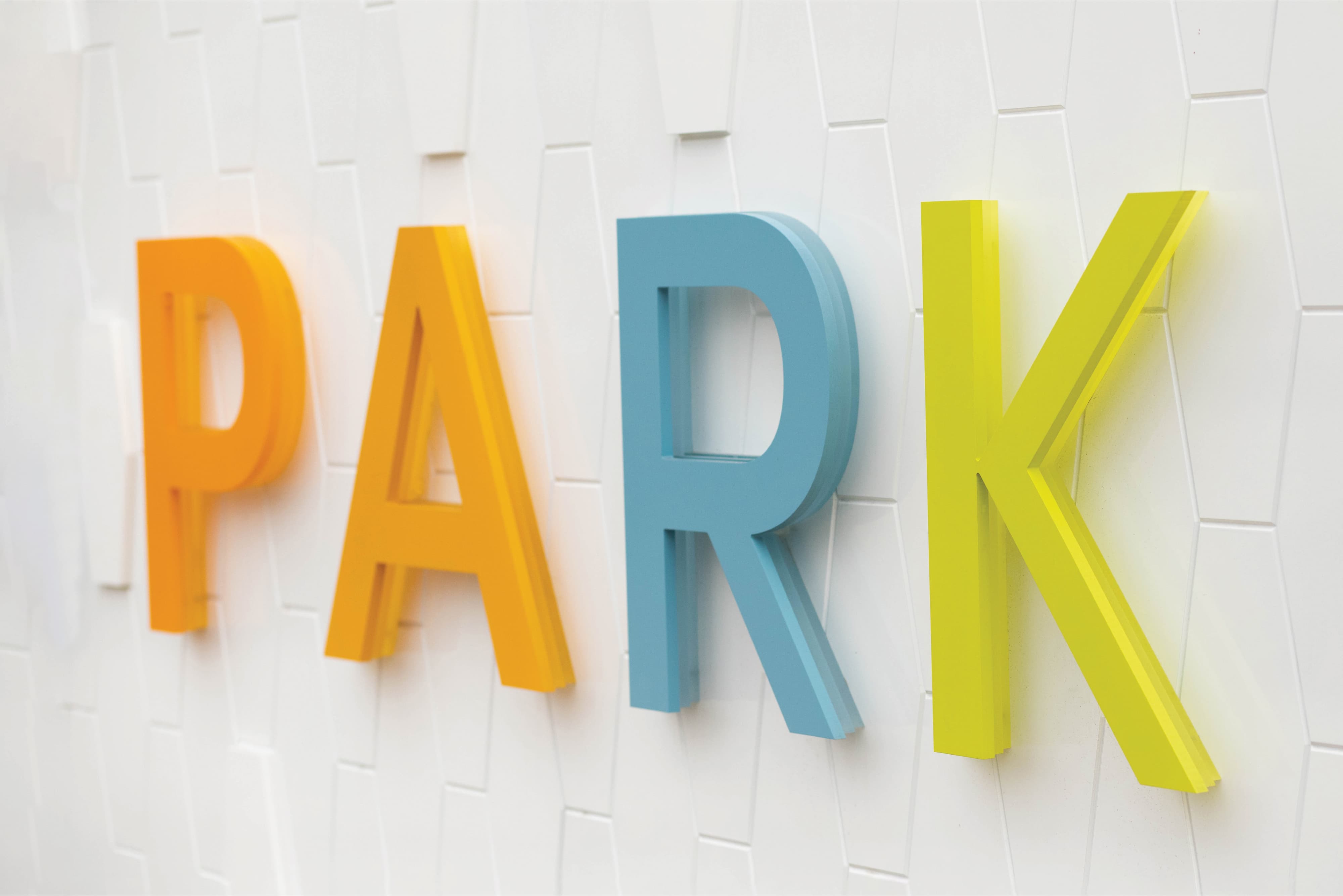Close-up detail of large-scale dimensional letters spelling "PARK" in alternating orange, teal, and lime green mounted on a textured white tile wall, echoing the Solis Park brand color palette — highlighting RSM Design's specialty graphics, experiential graphics and branded environments, and branding and visual identity services applied to identity signage within the residential community and park markets