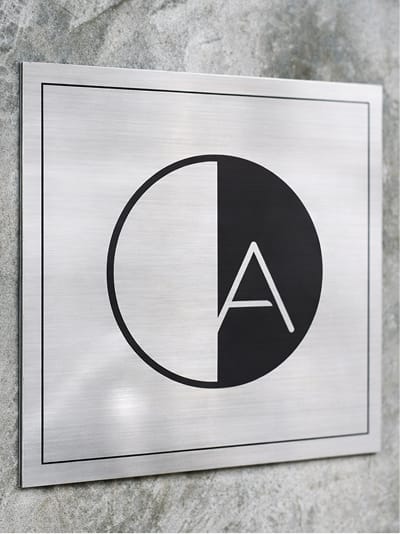 Brushed stainless steel wall-mounted identification plaque featuring the American Dream logo mark applied to a concrete surface at American Dream mall in East Rutherford, New Jersey — showcasing RSM Design's architectural graphics, specialty graphic design, and branded environments for a large-scale mixed-use retail and entertainment destination.