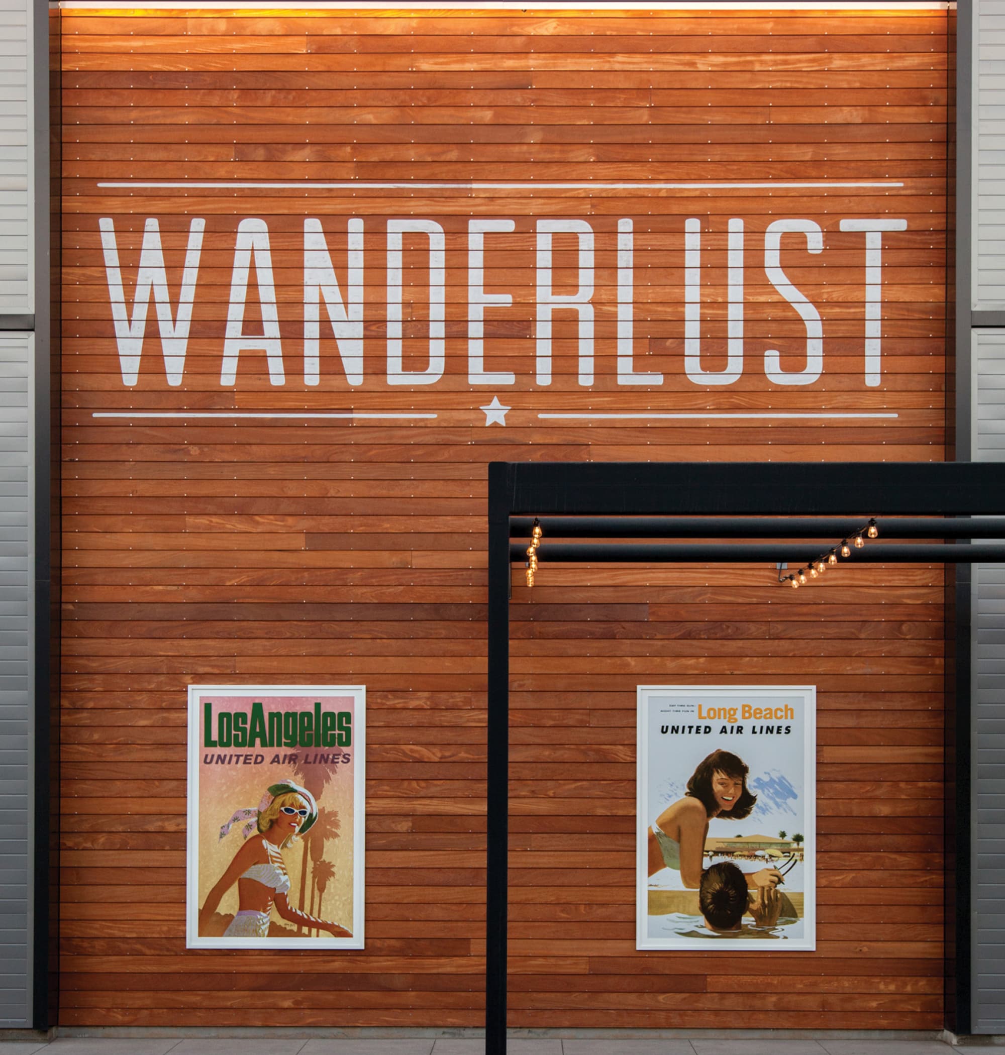 Wood slat wall painted with 'Wanderlust' in white stenciled letters between two horizontal lines and a star, above two vintage United Air Lines travel posters for Los Angeles and Long Beach, with string lights strung from a black frame to the right.