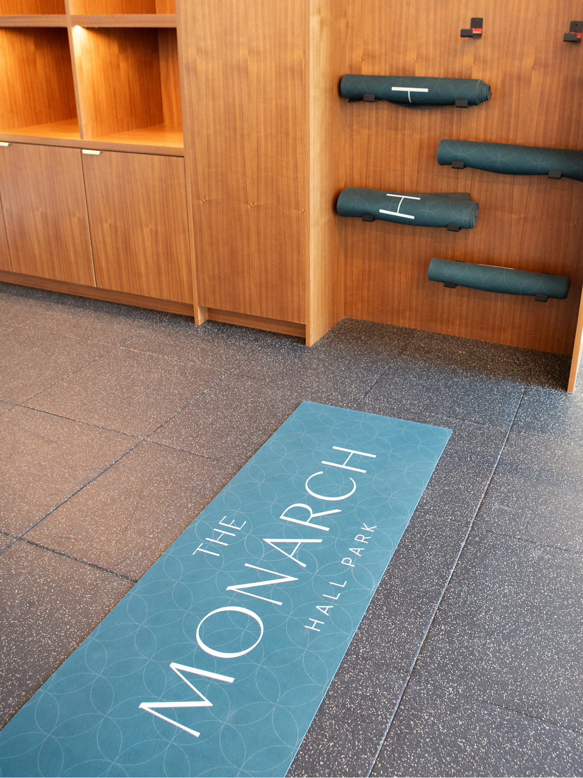 The Monarch at Hall Park branded fitness amenity merchandise — custom yoga mat with teal geometric pattern and Monarch Hall Park logo in yoga studio with wood-paneled mat storage wall, branded merchandise design by RSM Design
