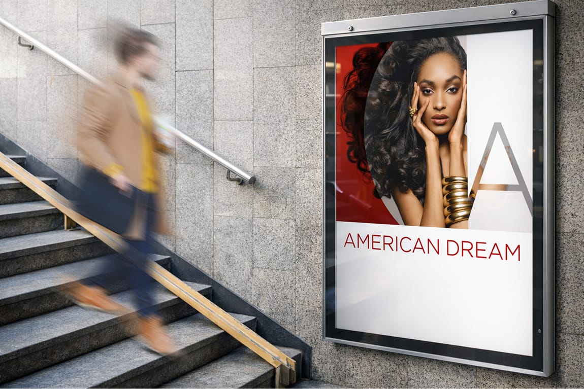 Illuminated transit poster display for American Dream mall in East Rutherford, New Jersey, featuring a fashion-forward brand campaign graphic with the American Dream logo mark and wordmark, mounted on a concrete stairwell wall with a pedestrian passing in motion — an example of RSM Design's print and digital collateral and branding and visual identity applied to a transit environment for a large-scale mixed-use retail and entertainment destination.
