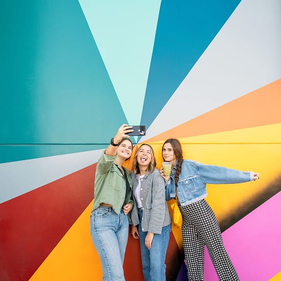 Three young women taking a selfie in front of the bold multicolored geometric mural on the exterior facade of Rodeo 39 Public Market in Stanton, CA.