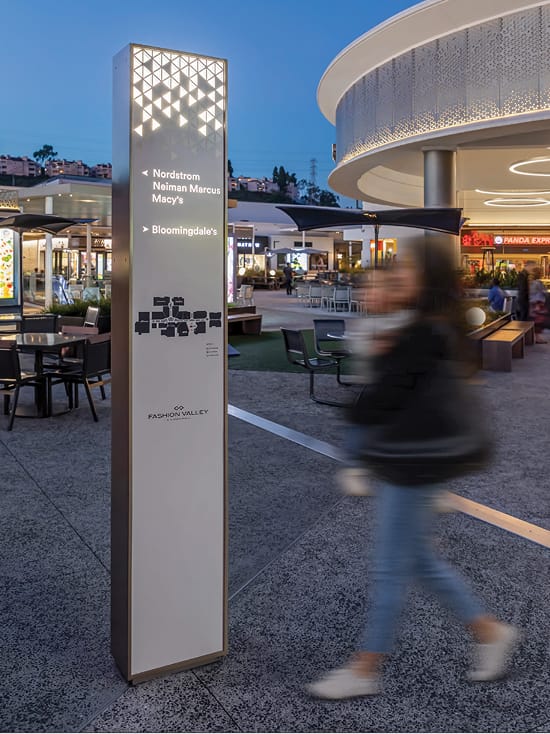Illuminated Fashion Valley wayfinding pylon at dusk directing shoppers to Nordstrom, Neiman Marcus, Macy's, and Bloomingdale's, with a motion-blurred pedestrian passing by and the mall's open-air dining and retail courtyard in the background.