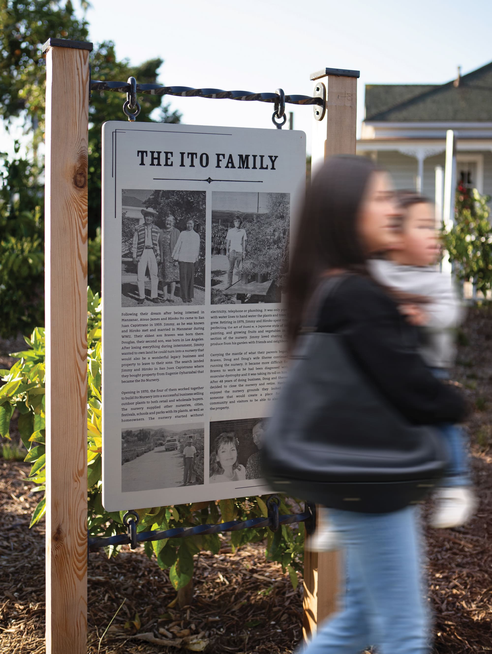 RSM Design interpretive heritage signage for River Street Marketplace, featuring an outdoor historical narrative panel telling the story of the Ito Family suspended from a wood and iron post structure amid a garden landscape, with a pedestrian passing in the foreground, demonstrating how environmental graphic design and placemaking can honor community history and cultural context to create meaningful, human-centered mixed-use and civic environments.