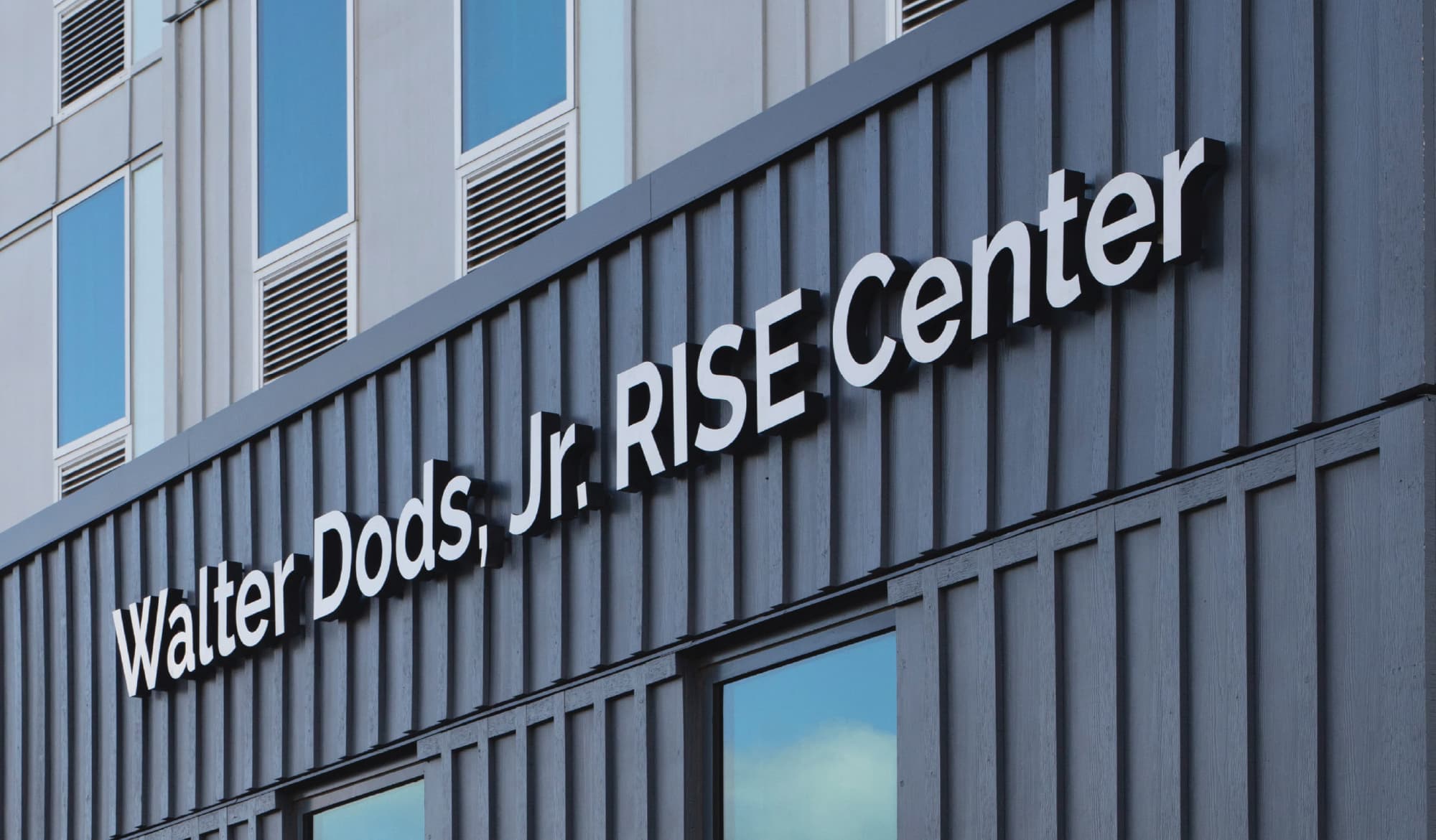 Angled view of dimensional white letters reading 'Walter Dods, Jr. RISE Center' mounted on a dark gray board-and-batten building facade.