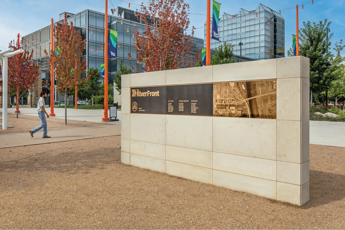 The RiverFront donor recognition wall in downtown Omaha, Nebraska, featuring a bronze dedication plaque with aerial map, limestone monument base, orange light towers, RiverFront branded banners, and modern glass office buildings in the background