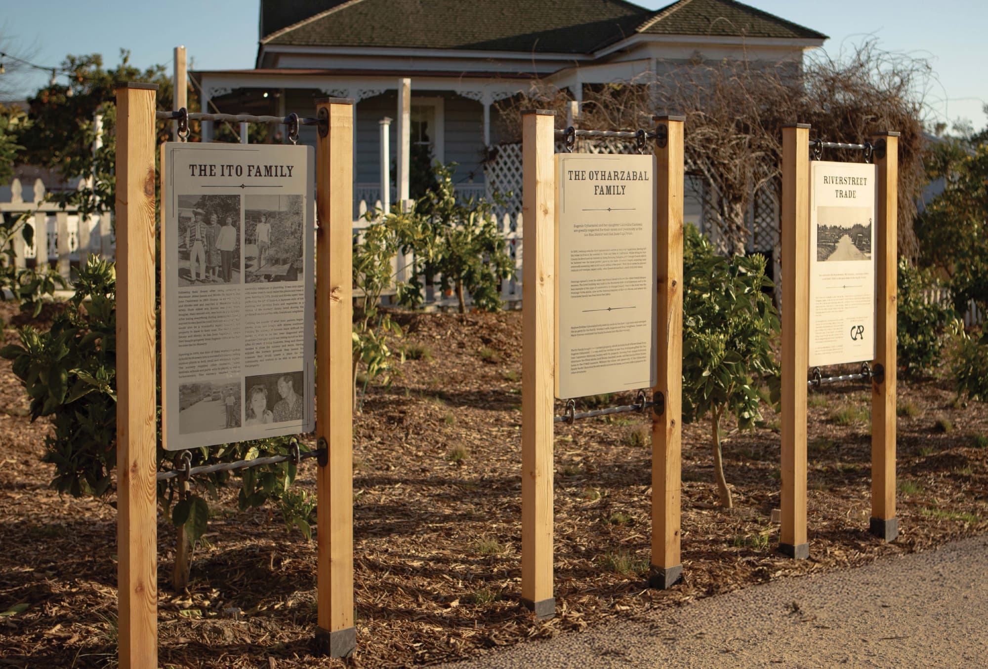 RSM Design interpretive signage and placemaking installation for River Street Marketplace, featuring a series of three outdoor heritage narrative panels — The Ito Family, The Oyharzabal Family, and River Street Trade — suspended from wood post structures in a garden landscape with a historic Victorian home in the background, illustrating how environmental graphic design, experiential graphics, and cultural storytelling unite to create meaningful, community-rooted mixed-use and civic environments.
