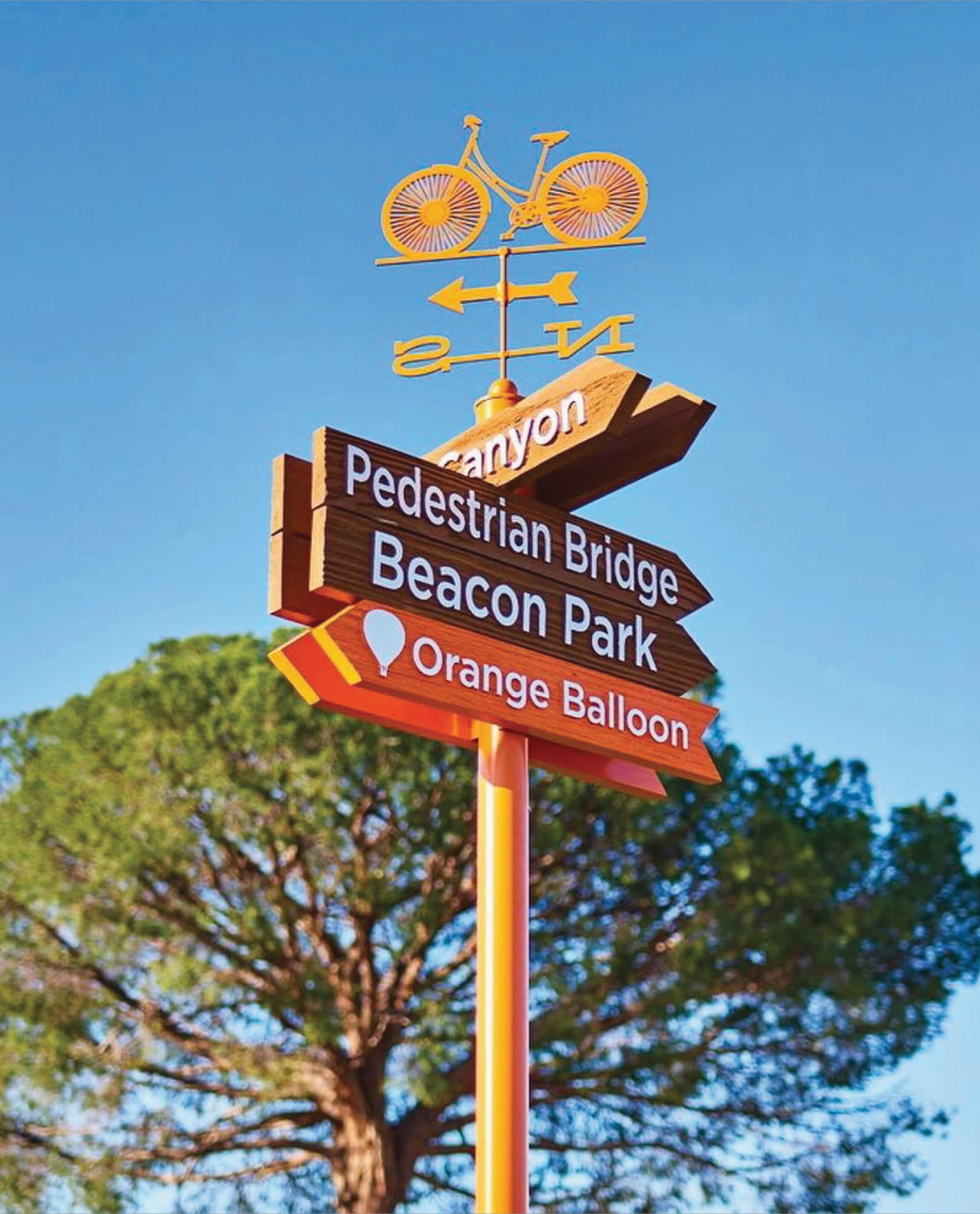 RSM Design pedestrian wayfinding signage and placemaking for FivePoint at Great Park Neighborhoods in Irvine, California, featuring a close-up of a whimsical multi-directional wood arrow sign cluster on a bright orange post topped with a gold bicycle weather vane, directing visitors to Canyon, Pedestrian Bridge, Beacon Park, and Orange Balloon destinations against a clear blue sky, showcasing how environmental graphic design, specialty graphics, experiential graphics, and wayfinding and signage design create joyful, human-centered navigation experiences that build community identity and a memorable sense of place for residential community and park markets.