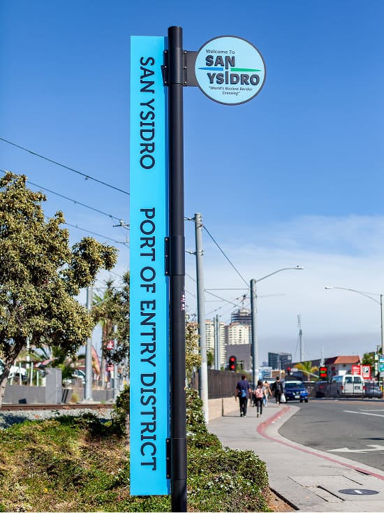 Blue vertical street sign reading 'San Ysidro Port of Entry District' with a circular 'Welcome to San Ysidro' emblem, positioned beside a curving road with trolley lines, pedestrians, and city buildings in the background.