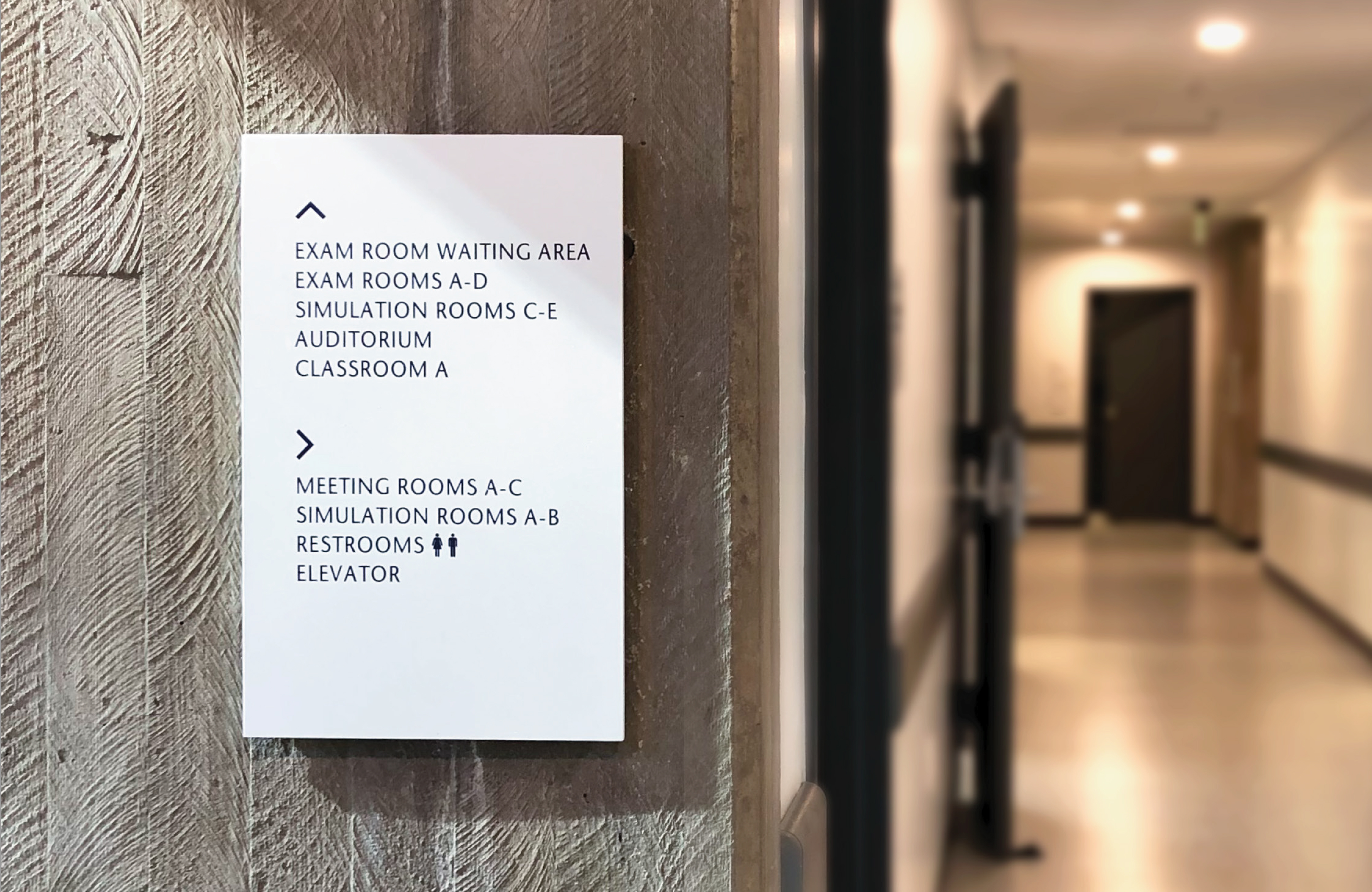 Interior wayfinding signage for a higher education campus building directing visitors to exam rooms, simulation labs, meeting rooms, and classrooms, illustrating environmental graphic design, experiential design, and integrated wayfinding systems.