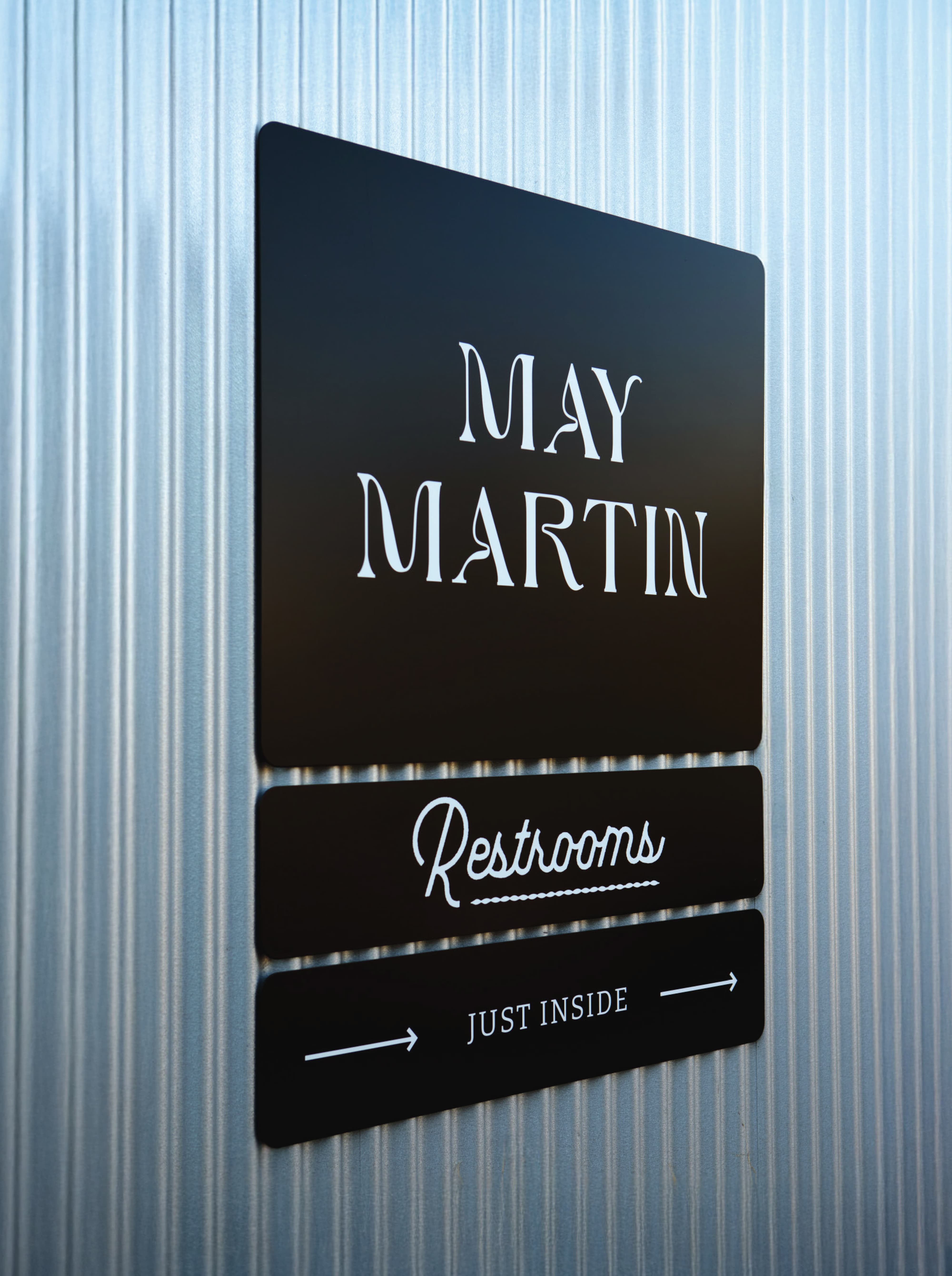 RSM Design wayfinding and signage design for River Street Marketplace, featuring a close-up of a stacked directional sign system for May Martin restaurant with branded restroom wayfinding panels in matte black mounted on a corrugated metal wall, demonstrating cohesive environmental graphic design and master sign program execution for food and beverage and mixed-use retail environments.
