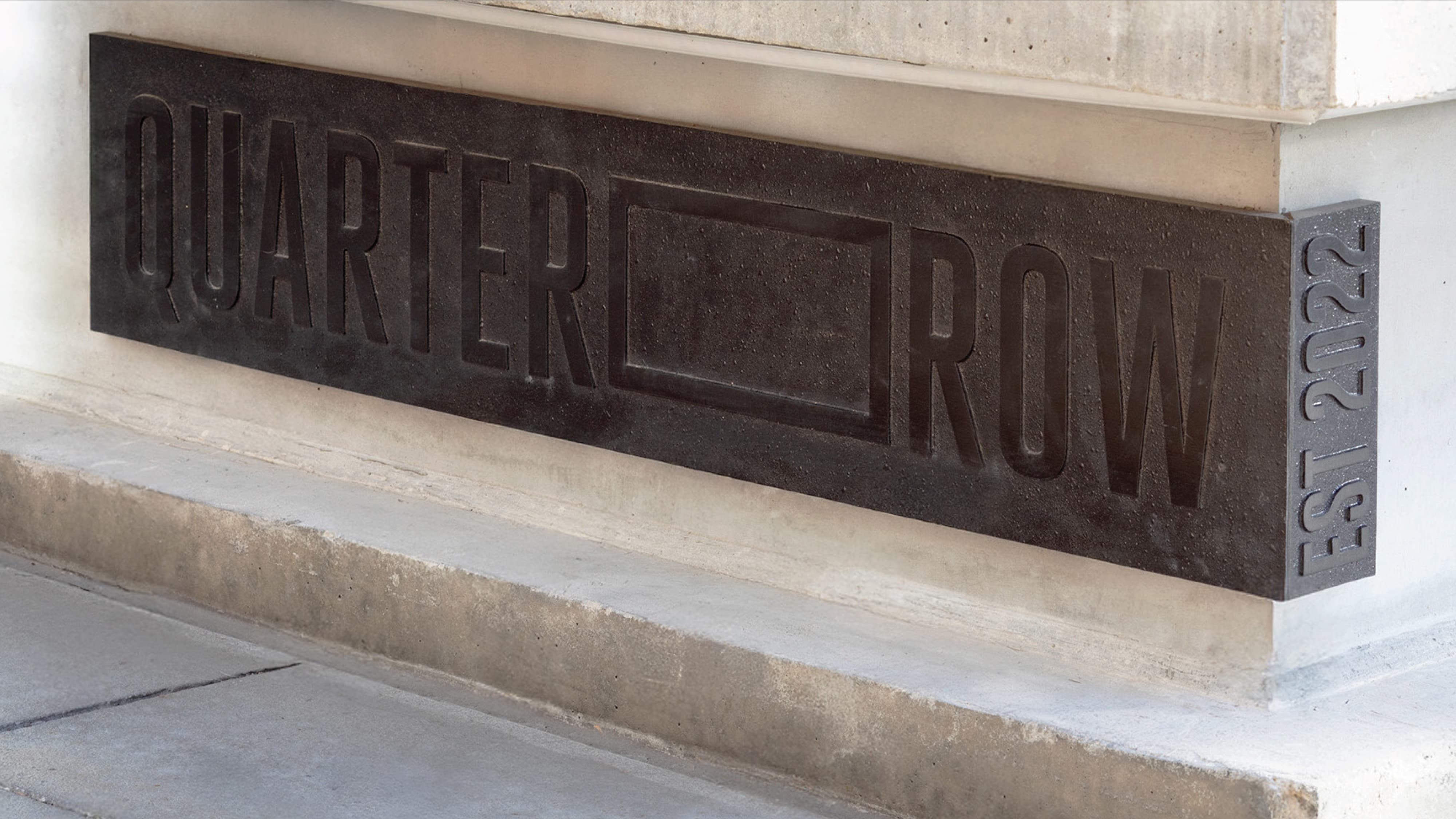 RSM Design architectural base signage and visual identity for Quarter Row at The West Quarter in Salt Lake City, featuring a close-up of a cast stone monument panel with deeply routed sans-serif dimensional lettering and an established date of 2022 inscribed vertically on the return edge, set flush against a concrete building plinth, illustrating how environmental graphic design and specialty graphics treat identity signage as a permanent architectural element — grounding place, communicating legacy, and creating a lasting branded environment for mixed-use, residential community, and urban streetscape markets.