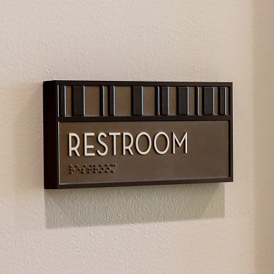 Bronze restroom sign with a striped pattern header, braille text, and 'Restroom' in white lettering, mounted on a textured wall.