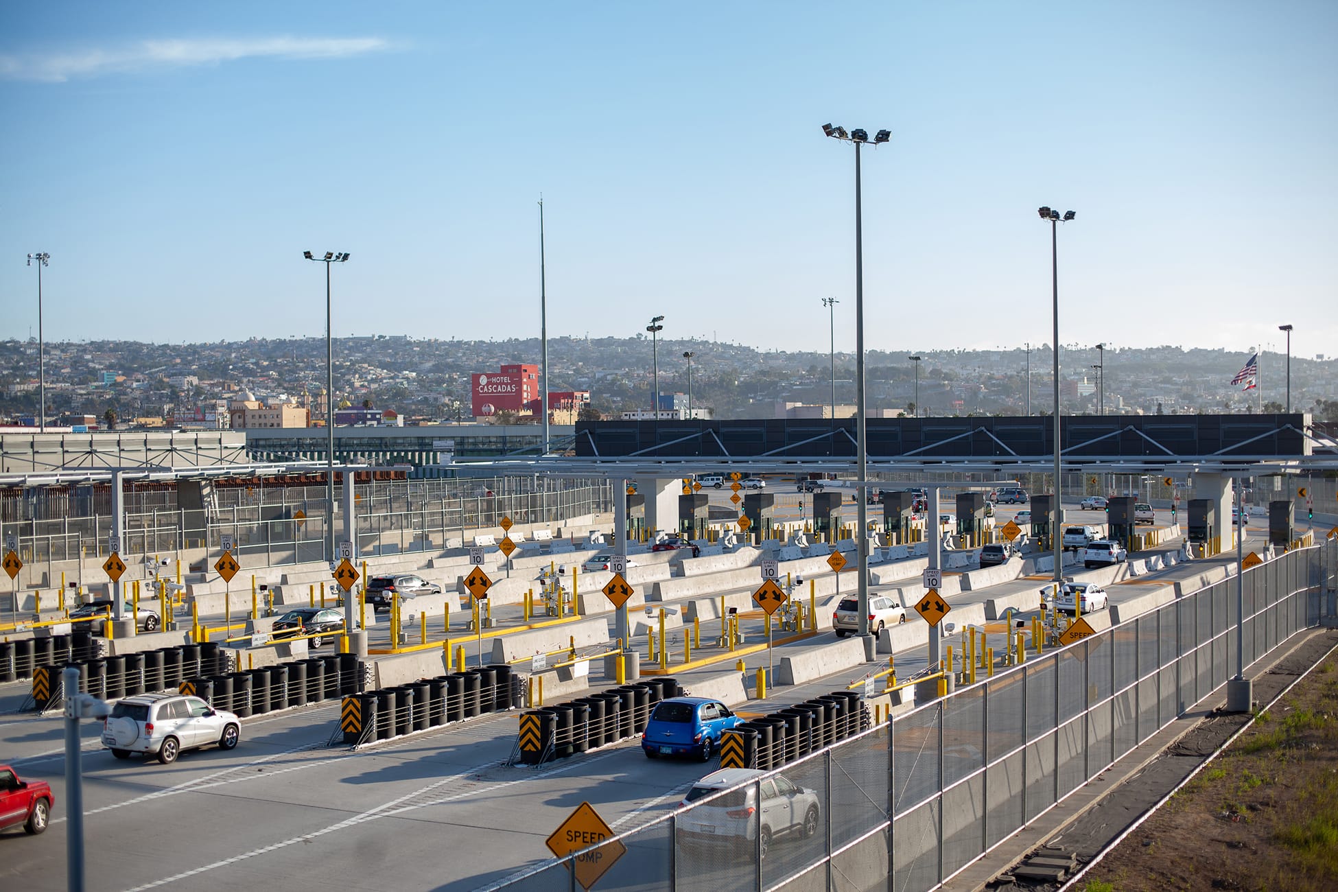 Wide view of vehicle inspection lanes at the San Ysidro Port of Entry, with cars passing through concrete barriers and speed limit signs beneath tall light poles, the Tijuana cityscape and 'Hotel Cascadas' sign visible in the hazy background.