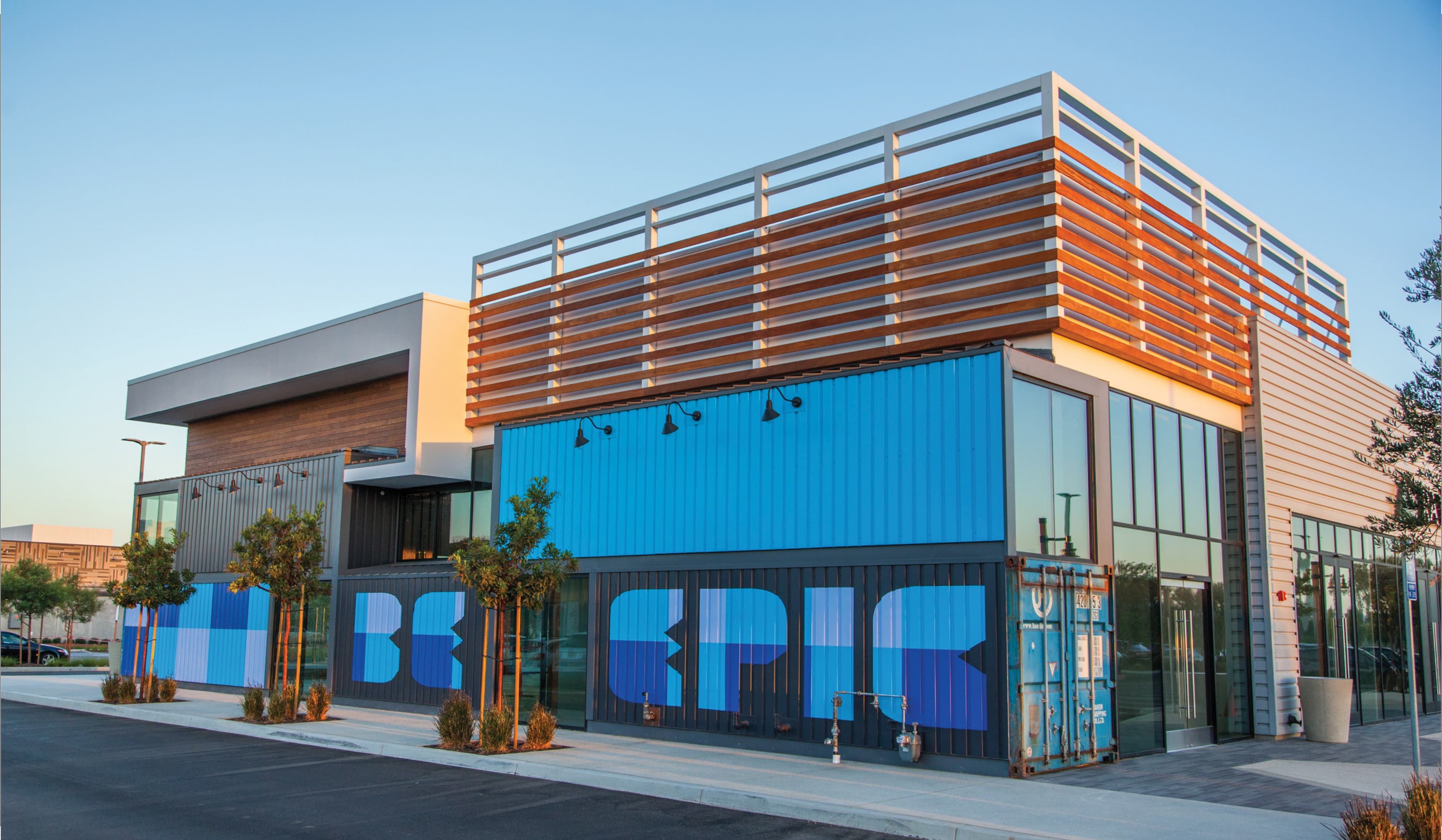 Modern retail building constructed from stacked blue shipping containers with a wood slat rooftop screen, painted with a large 'Be Epic' mural in shades of blue across the storefronts, young trees lining the sidewalk at golden hour.