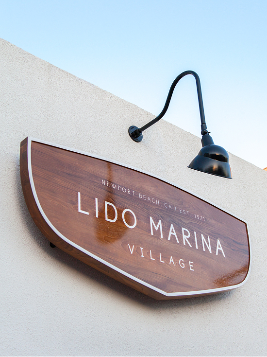 Close-up of Lido Marina Village wood blade sign shaped like a boat hull with white typography reading "Newport Beach, CA Est. 1971" and black gooseneck wall light, Newport Beach, California