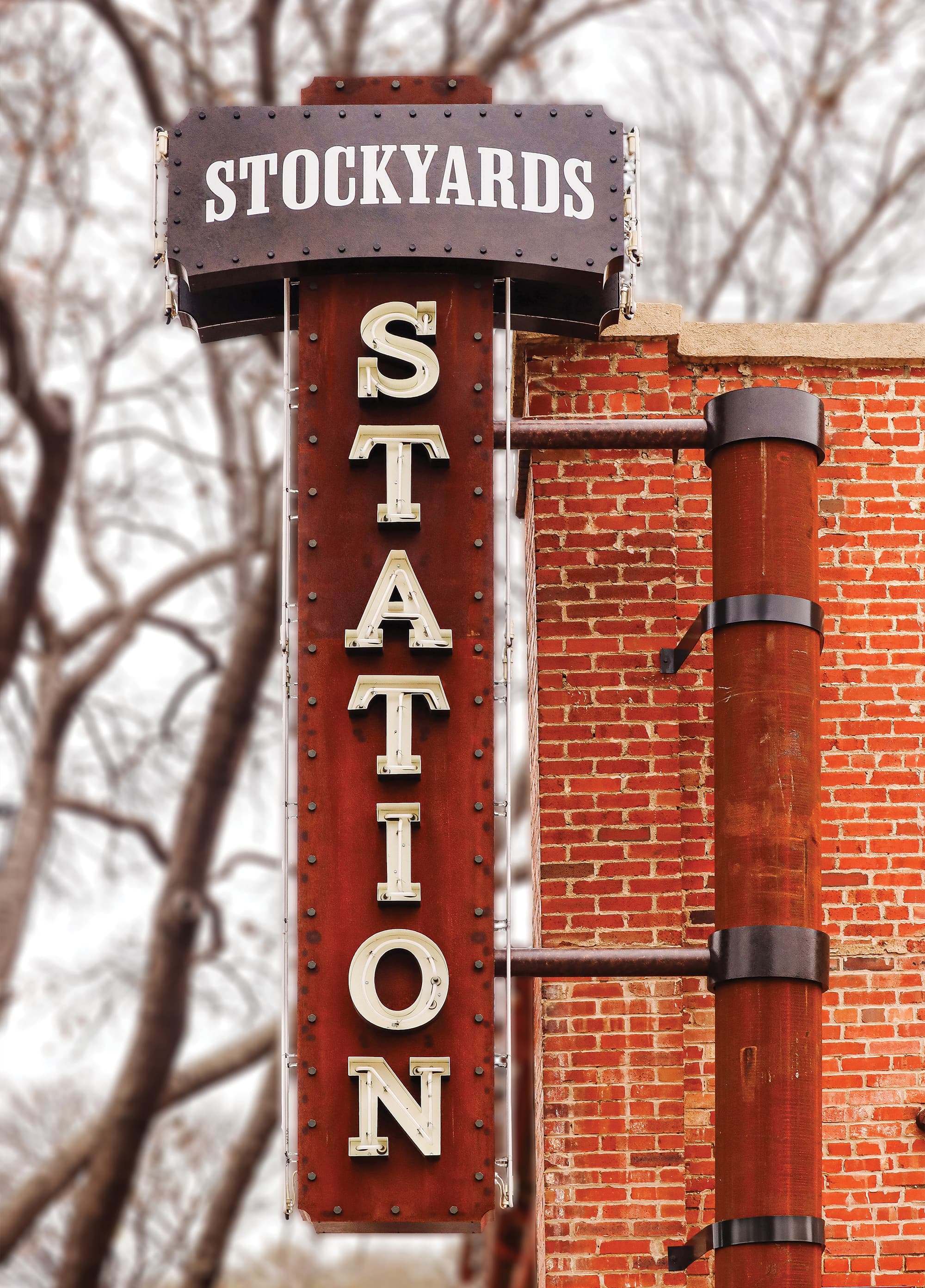 A cantilever sign attached to a pole, with reinforcement anchors to a brick wall. Text on the sign reads “Stockyards Station” in all capital letters in an antique slab serif. The sign face is made of two sheets of weathered, rust-brown sheet metal bordered with rivets, arranged to resemble a sledgehammer.