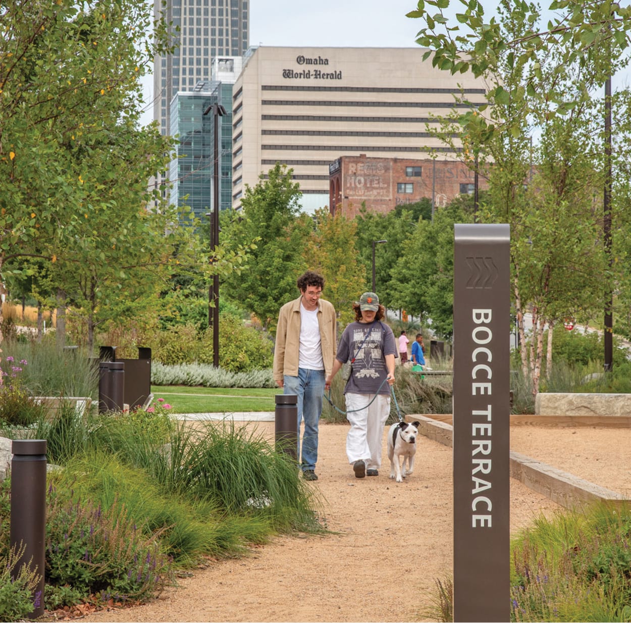 Couple walking their dog along the Bocce Terrace pathway at Lewis and Clark Landing riverfront park in downtown Omaha, Nebraska, with lush landscaping, native plantings, and the Omaha World-Herald building visible in the background