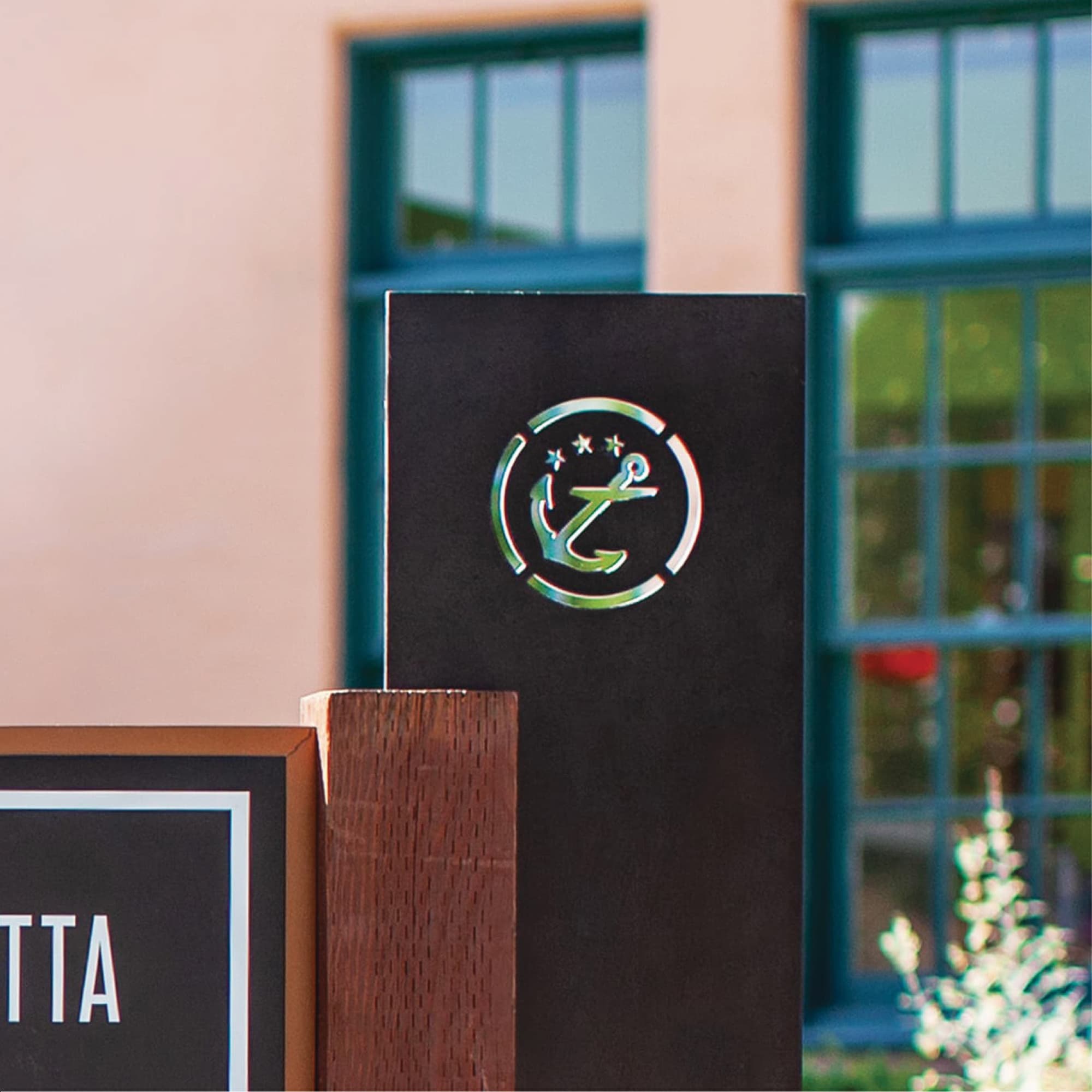 Close-up of a black wayfinding sign with the Liberty Station anchor and stars emblem cut out and lit with a green internal glow, positioned beside a wooden post and partial black sign reading 'TTA,' with teal-framed windows in the background.