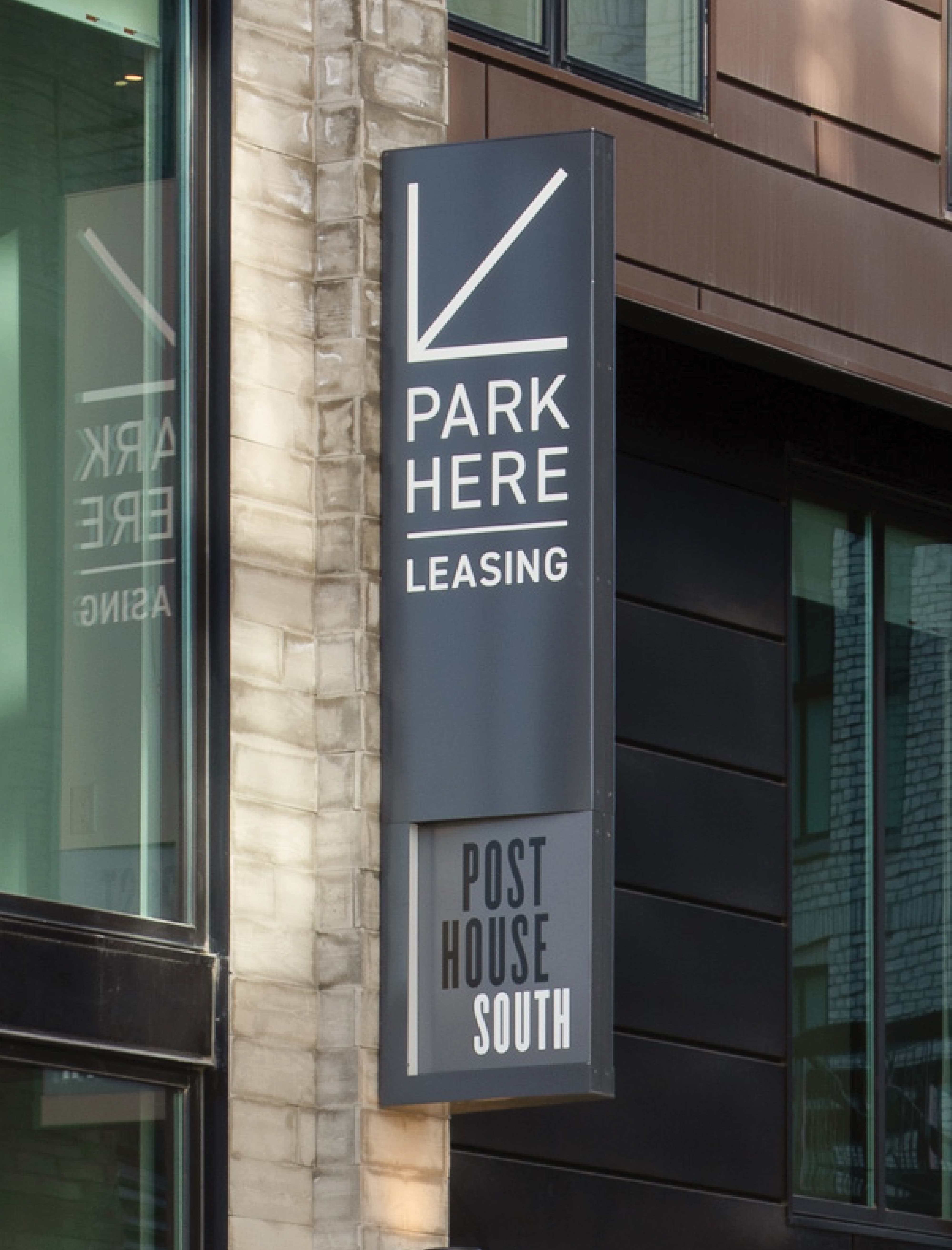 Exterior projecting blade sign mounted on a stone column at Post House South at Post District in Salt Lake City, displaying a directional arrow graphic with 'Park Here — Leasing' messaging above the Post House South sub-brand identification — an example of RSM Design's wayfinding signage design and environmental graphic design for a mixed-use residential community.