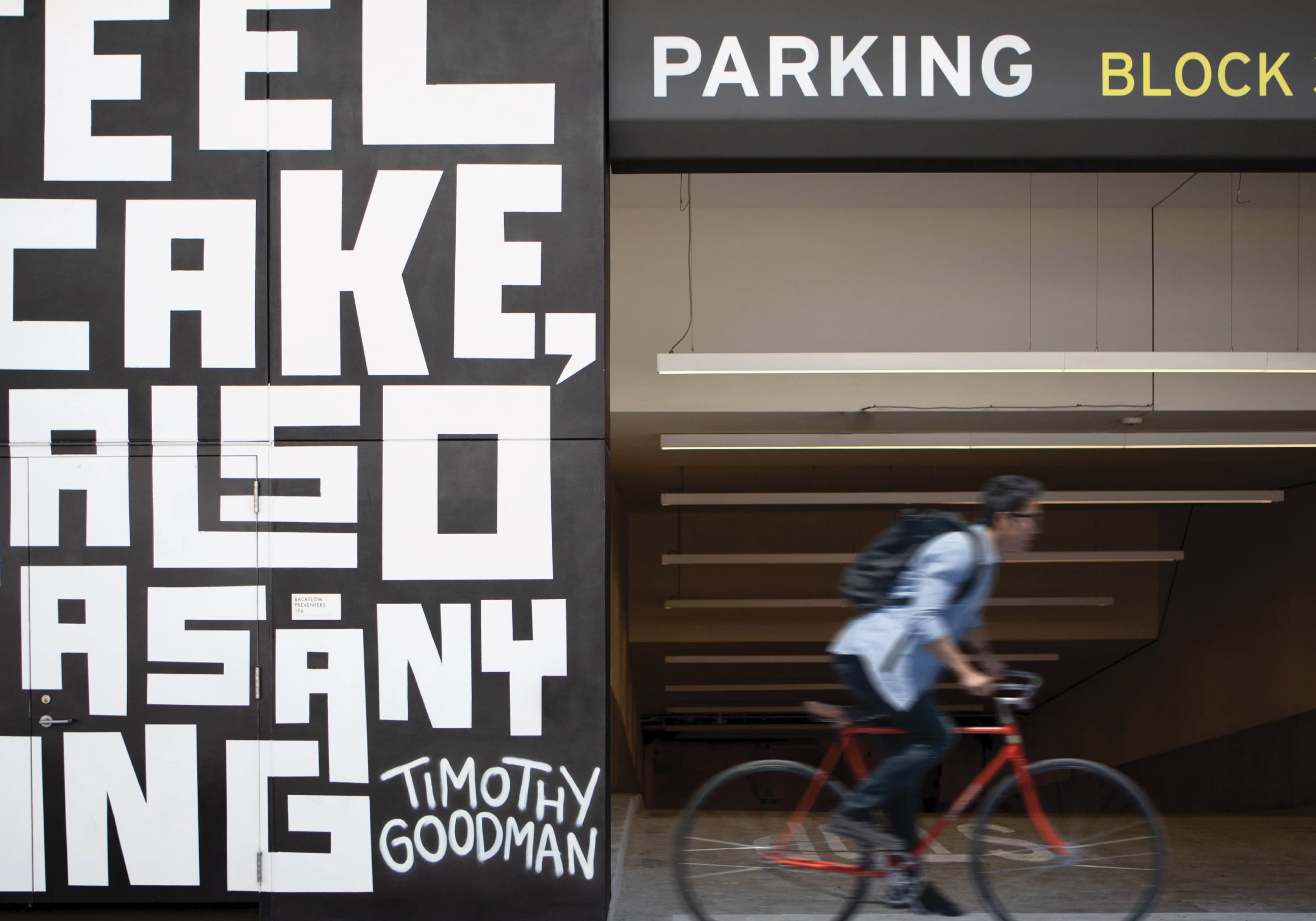 Bold black and white typographic mural reading partial phrases like 'eel, ake, also, as any, ing' signed by artist Timothy Goodman, beside a parking garage entrance with a 'Parking Block' sign, as a man on a red bicycle rides past.
