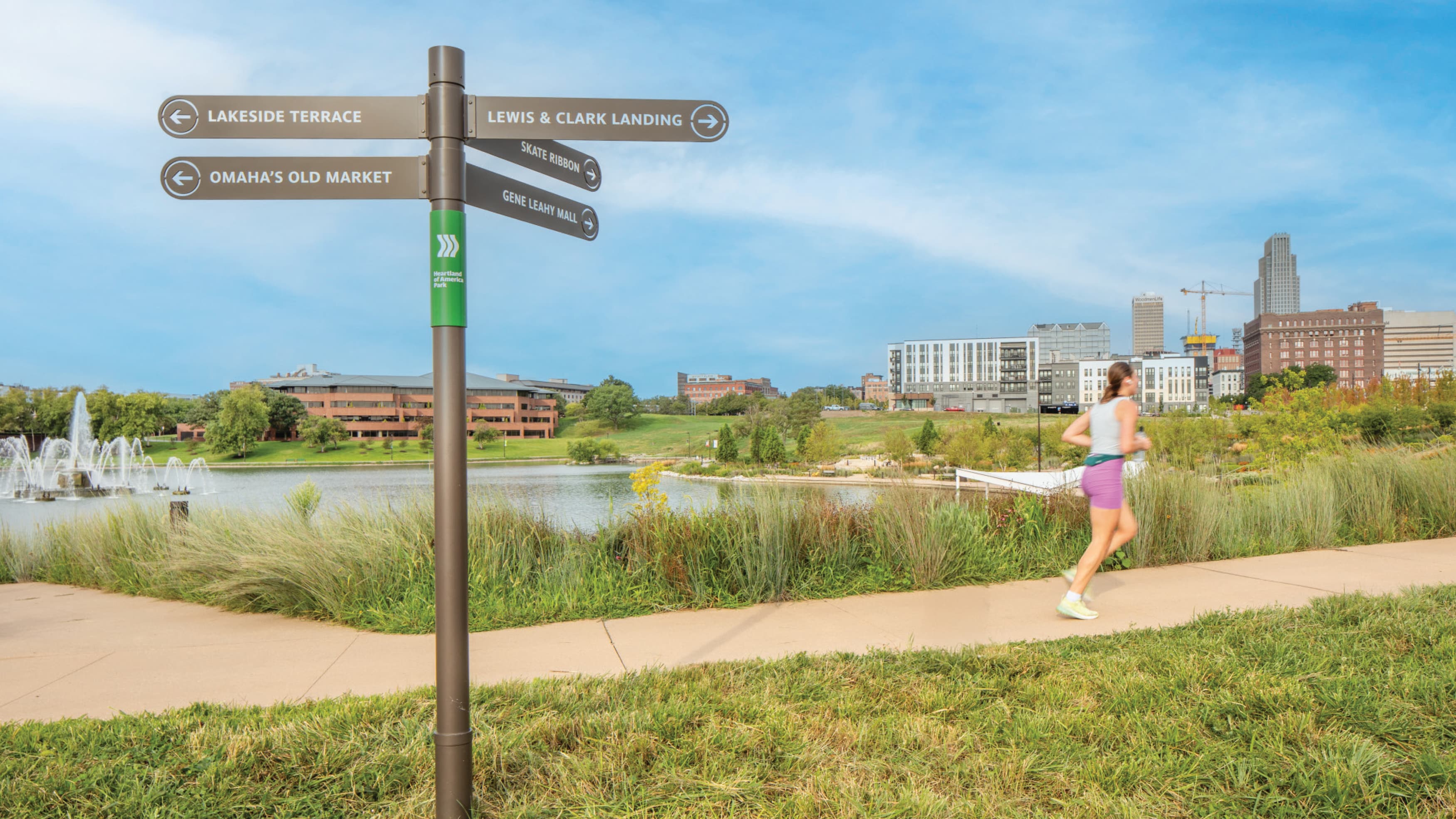 
The image shows wayfinding signage designed by RSM Design in a park setting, with a lake and city skyline in the background. The signage provides directions to various local attractions, such as Lakeside Terrace, Lewis & Clark Landing, Omaha's Old Market, Skate Ribbon, and Gene Leahy Mall. A woman in athletic wear is jogging on a path near the signage, highlighting the park's recreational use. The environment is lush with green grass and shrubs, and the weather appears clear and sunny, contributing to a vibrant, active, and inviting urban park atmosphere. This wayfinding signage, created by RSM Design, helps visitors navigate the park easily and enhances their overall experience.