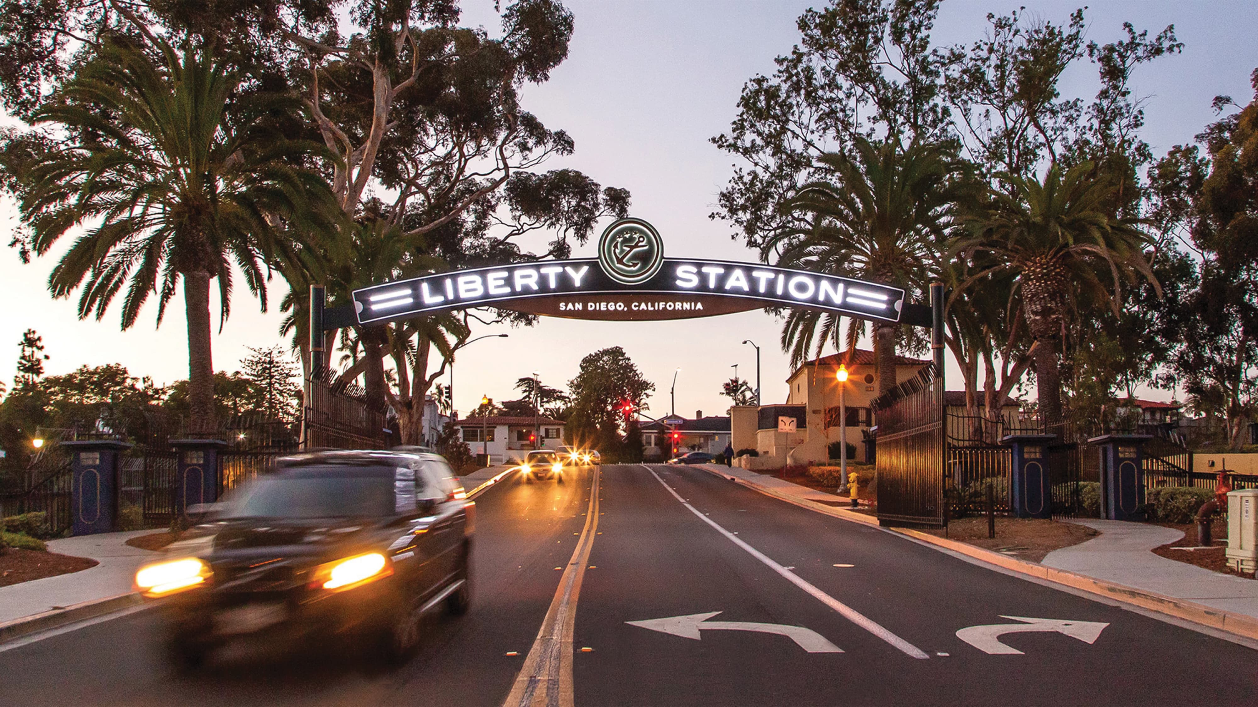 Illuminated archway sign reading 'Liberty Station, San Diego, California' with an anchor emblem, spanning a street lined with palm trees and wrought-iron gates, with cars driving beneath at dusk.