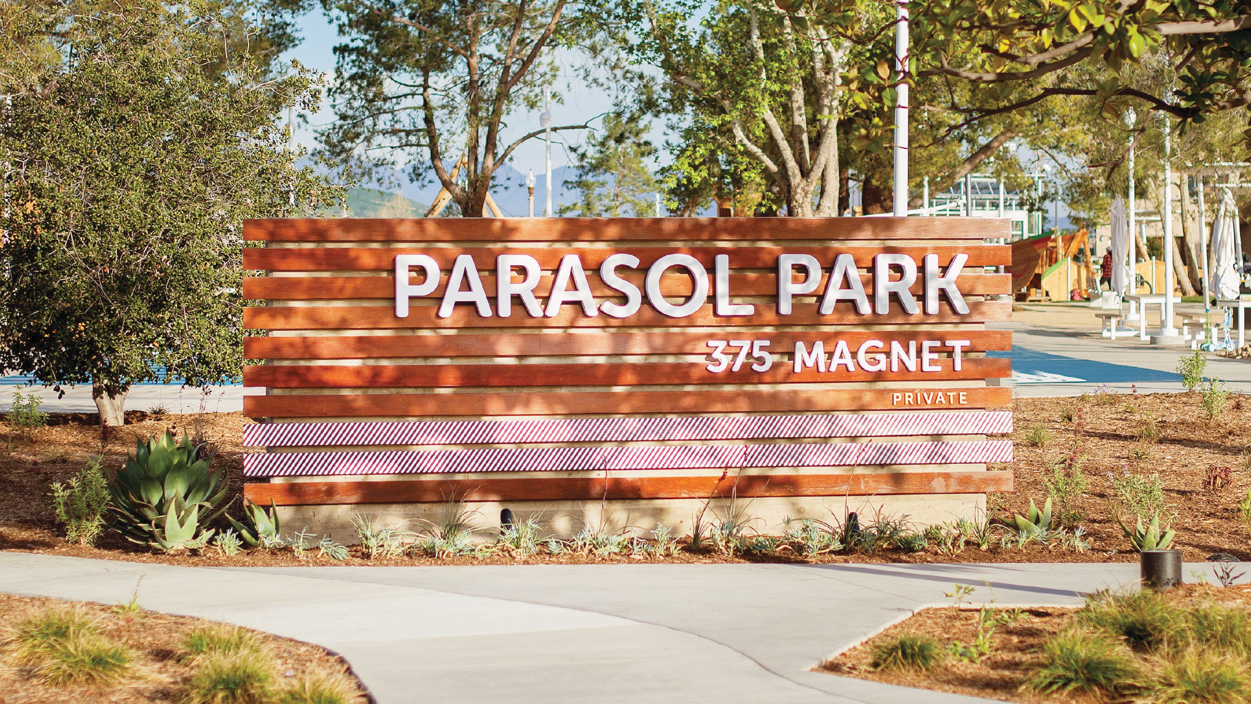 RSM Design monument signage and placemaking for Parasol Park at 375 Magnet, featuring a close-up of a warm horizontal wood slat identity sign with dimensional white lettering and a playful red and white diagonal stripe accent band, set within a drought-tolerant landscaped planting bed with agave and succulents, with a playground and mature tree canopy visible in the background, demonstrating how environmental graphic design, landmark identity signage, wayfinding and signage design, and branded environments create a welcoming, character-rich sense of arrival and place for residential community, park, and mixed-use waterfront markets.