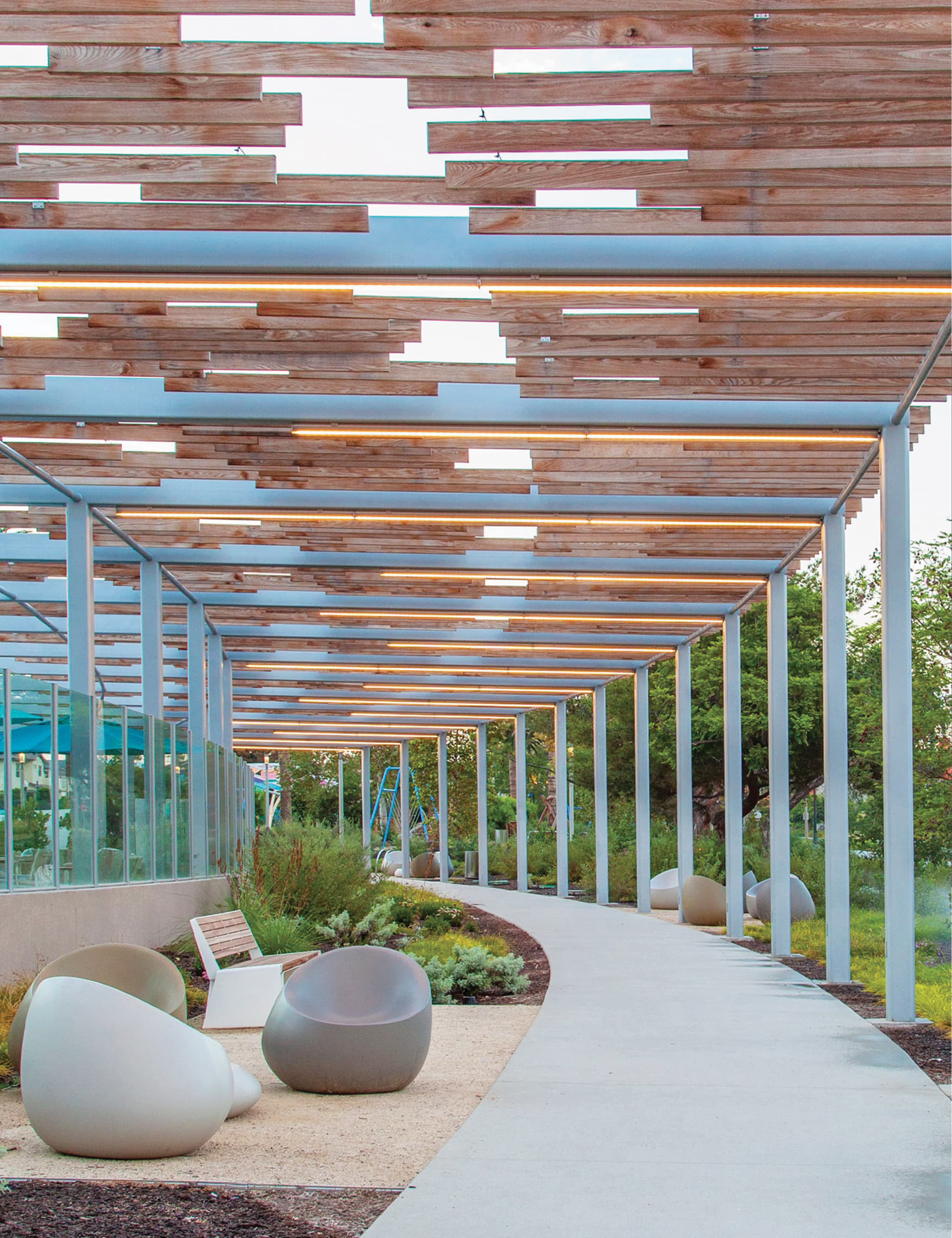 Covered walkway with a staggered wood slat canopy and integrated linear lighting, running alongside a glass-walled building, with a curved path, egg-shaped stone seating, a bench, and landscaped greenery beside it.