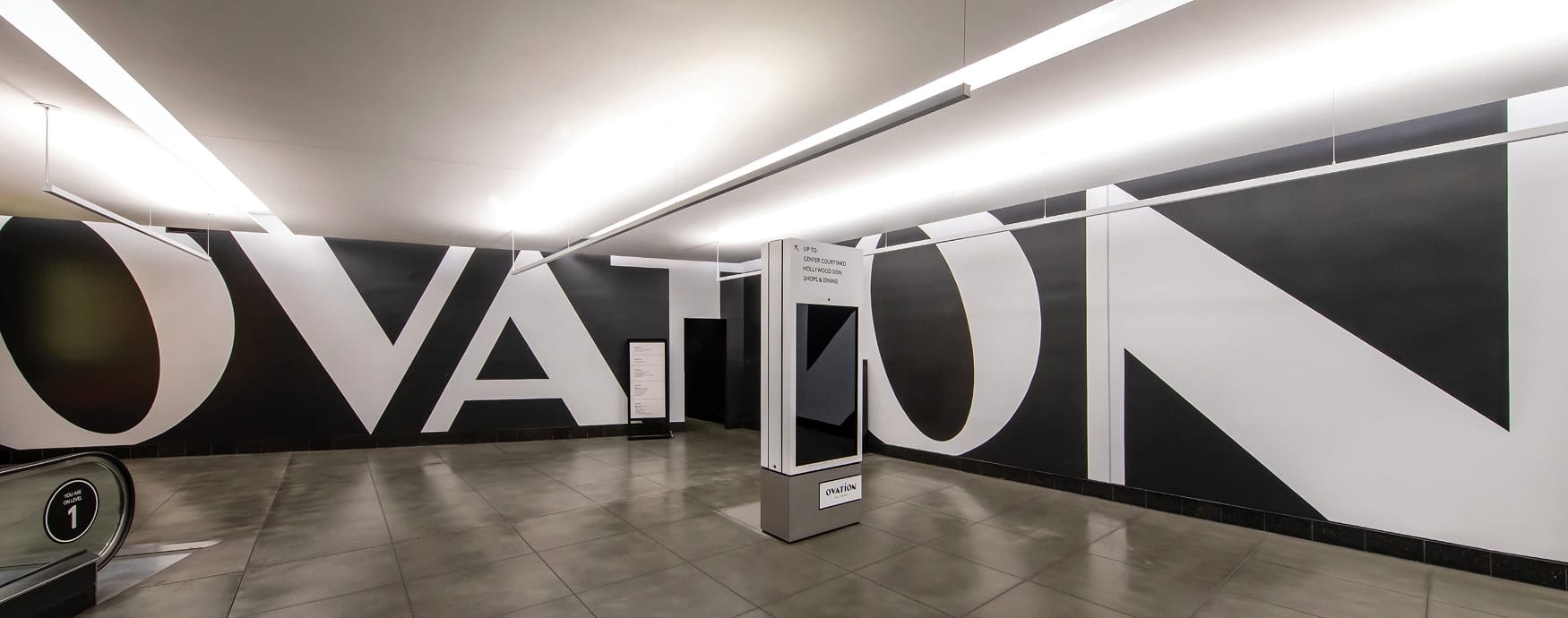 Ovation Hollywood Level 1 parking garage interior with dramatic oversized black and white "OVATION" typographic wall mural, digital wayfinding kiosk directing visitors to Center Courtyard, Hollywood Sign, shops and dining
