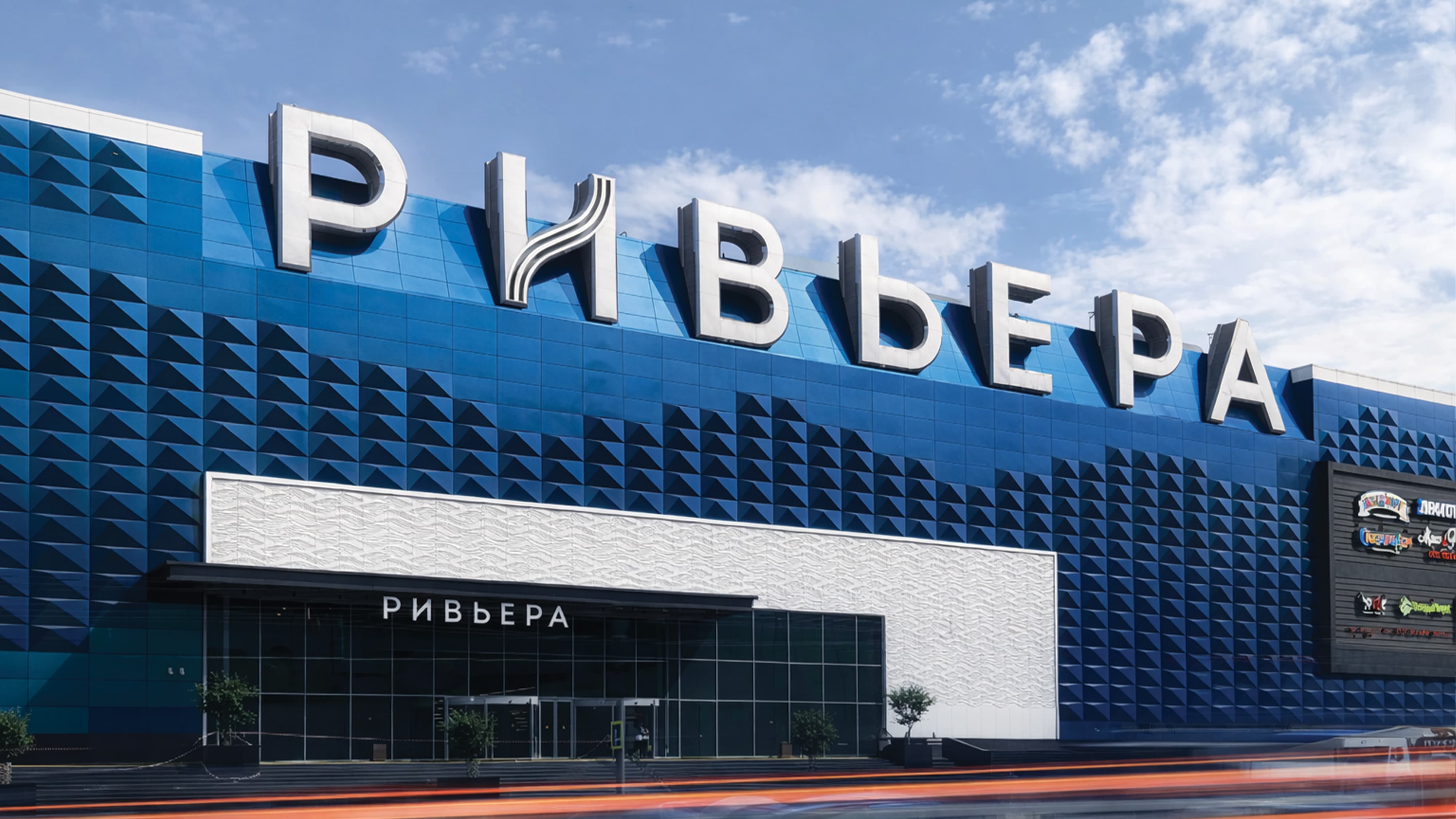 Exterior street-level view of Riviera international retail and mixed-use destination in Moscow, Russia, featuring large-scale rooftop dimensional Cyrillic building identification signage, entry canopy signage, tenant directory sign panel, and geometric triangulated blue building facade — RSM Design branding and visual identity, wayfinding and signage design, master sign programs, specialty graphics, and environmental graphic design for retail, mixed-use, streetscape, and international destinations.