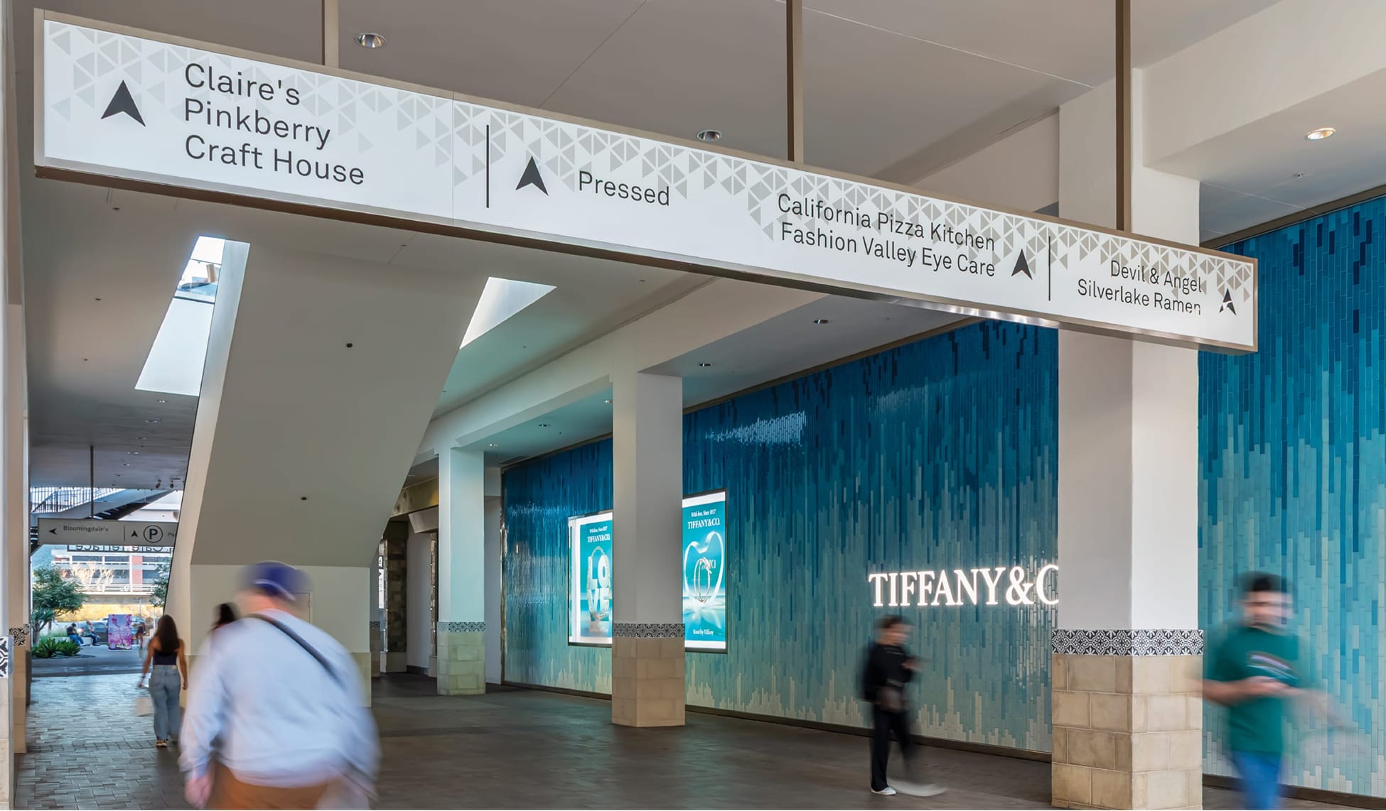 Interior corridor at Fashion Valley Mall showing an overhead blade wayfinding sign directing shoppers to Claire's, Pinkberry, Craft House, Pressed, California Pizza Kitchen, Devil & Angel, and Silverlake Ramen, with the Tiffany & Co. storefront's signature blue tile facade visible beyond.