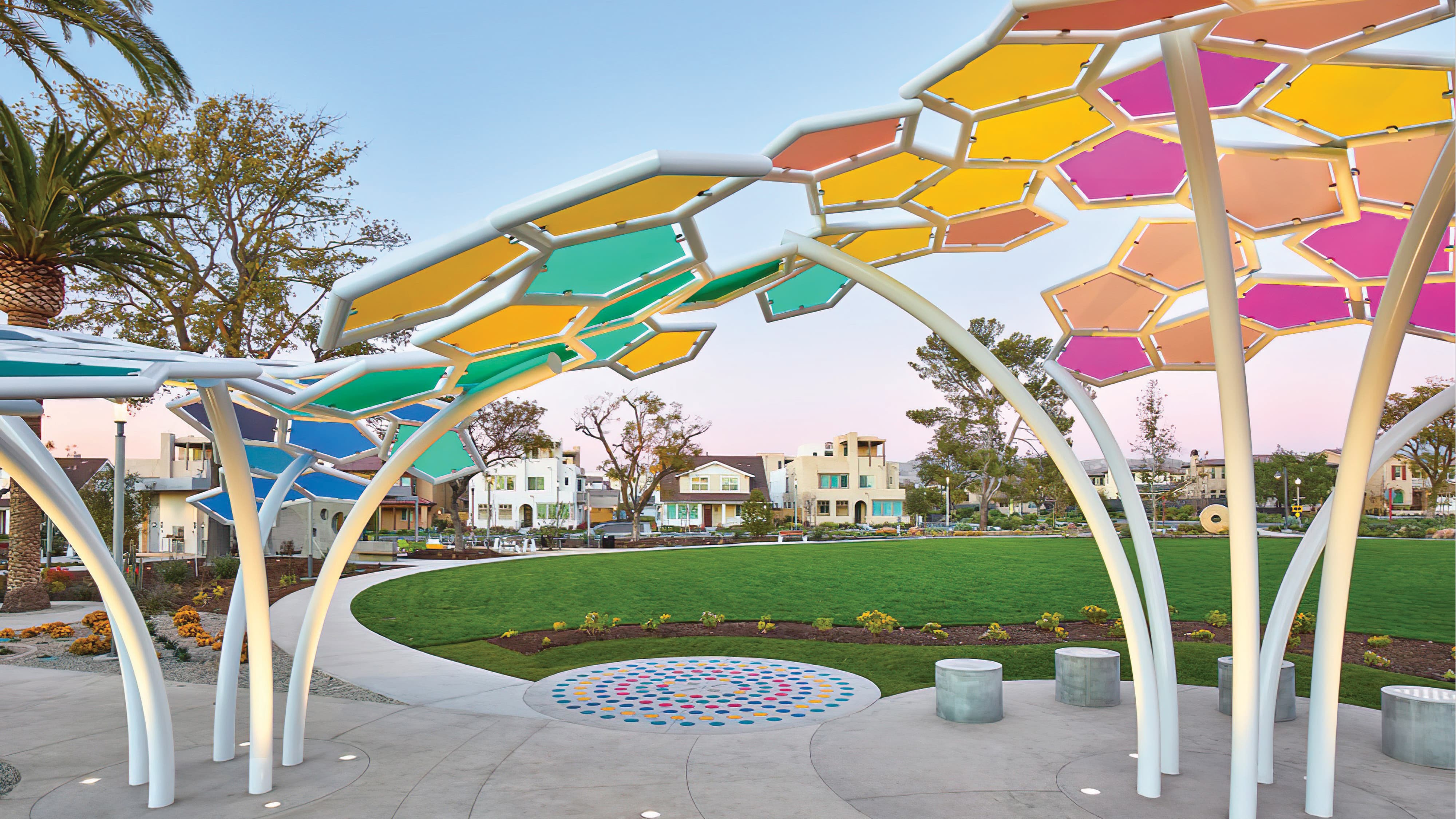 Sculptural park pavilion with slender white columns supporting a canopy of multicolored hexagonal panels in teal, gold, pink, and blue, shading a circular plaza with a mosaic dot pattern and stone seating stools, overlooking an open lawn and residential neighborhood at dusk.