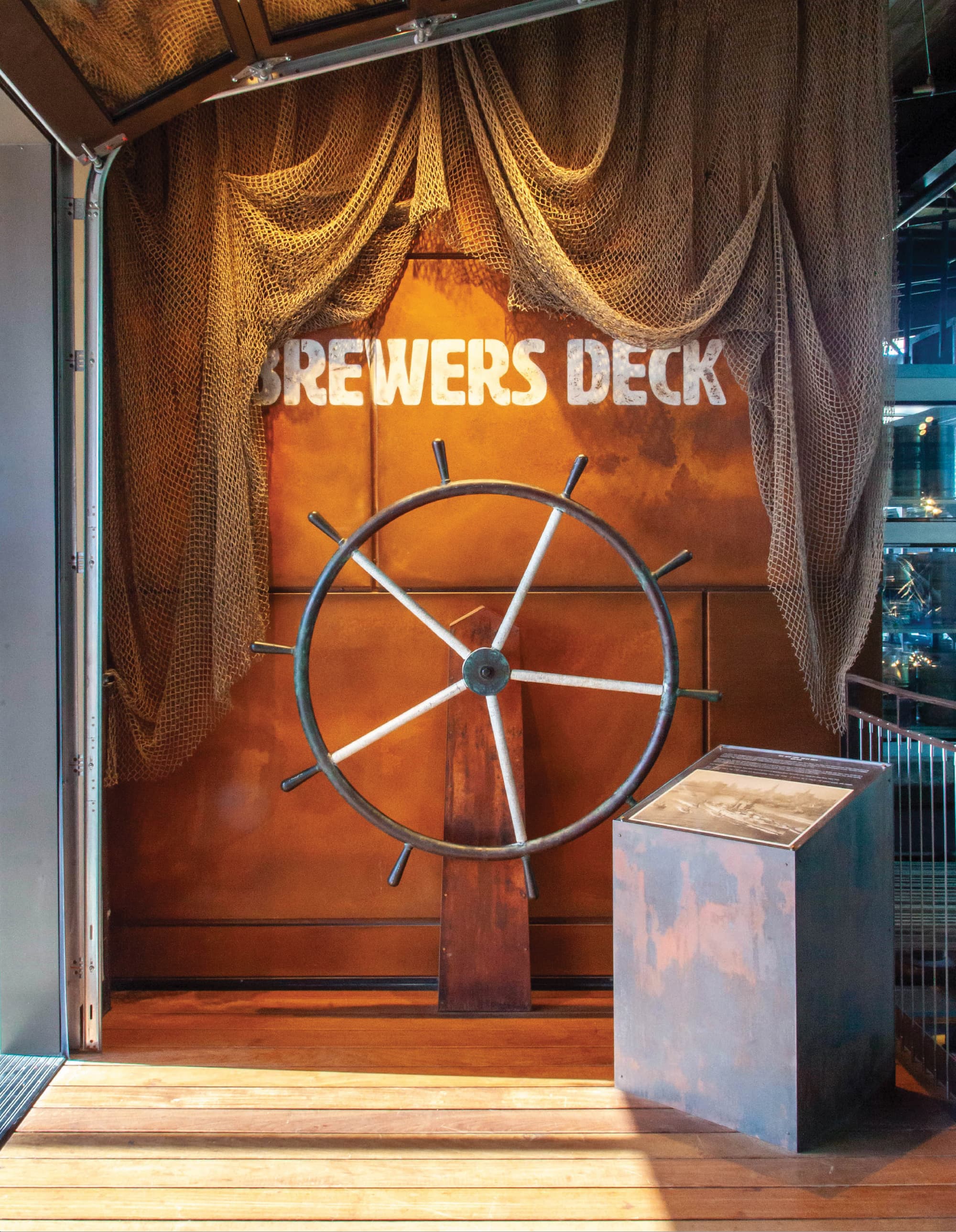 A dramatic nautical-themed arrival vignette at Crafthouse Brewers Deck featuring a large antique ship's wheel centered against a weathered copper wall with stenciled "Brewers Deck" lettering, draped fishing net, warm wood decking, and an interpretive panel — exemplifying RSM Design's placemaking, experiential graphics and branded environments, and specialty graphics services that create immersive, story-driven moments within the food and beverage and mixed-use markets