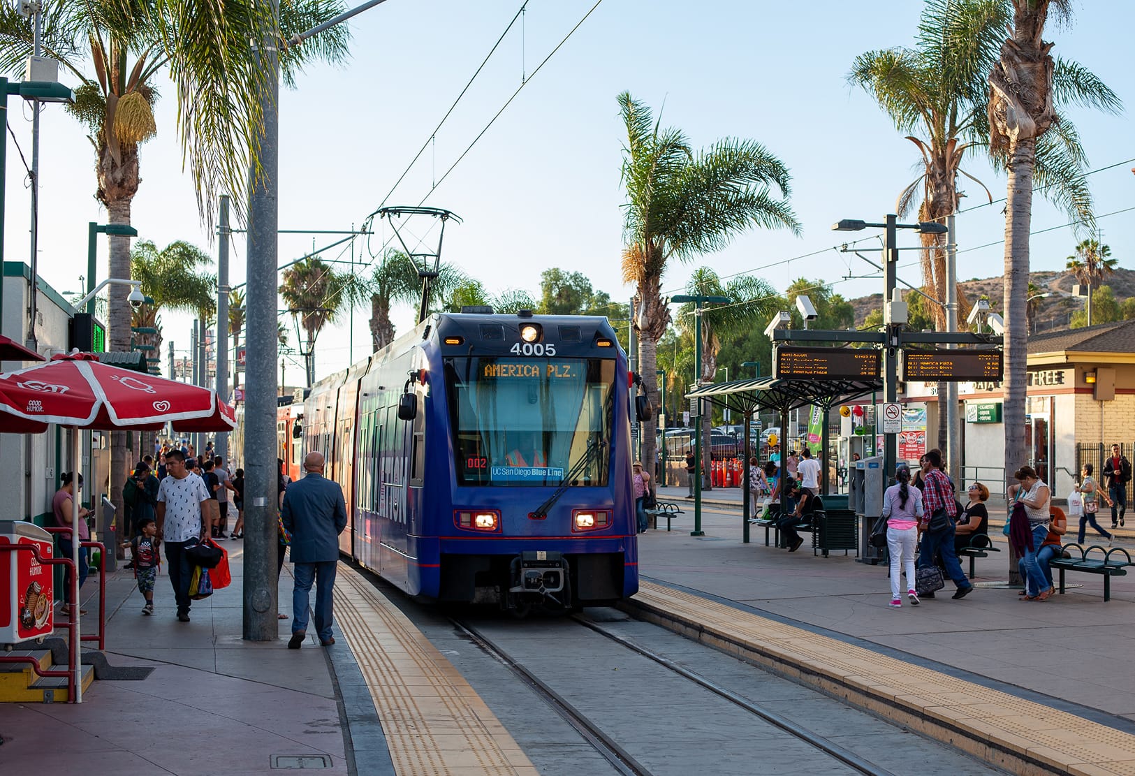 Blue San Diego Trolley UC San Diego Blue Line train numbered 4005 arriving at the San Ysidro station platform, with waiting passengers, vendor carts, palm trees, and hills in the background.
