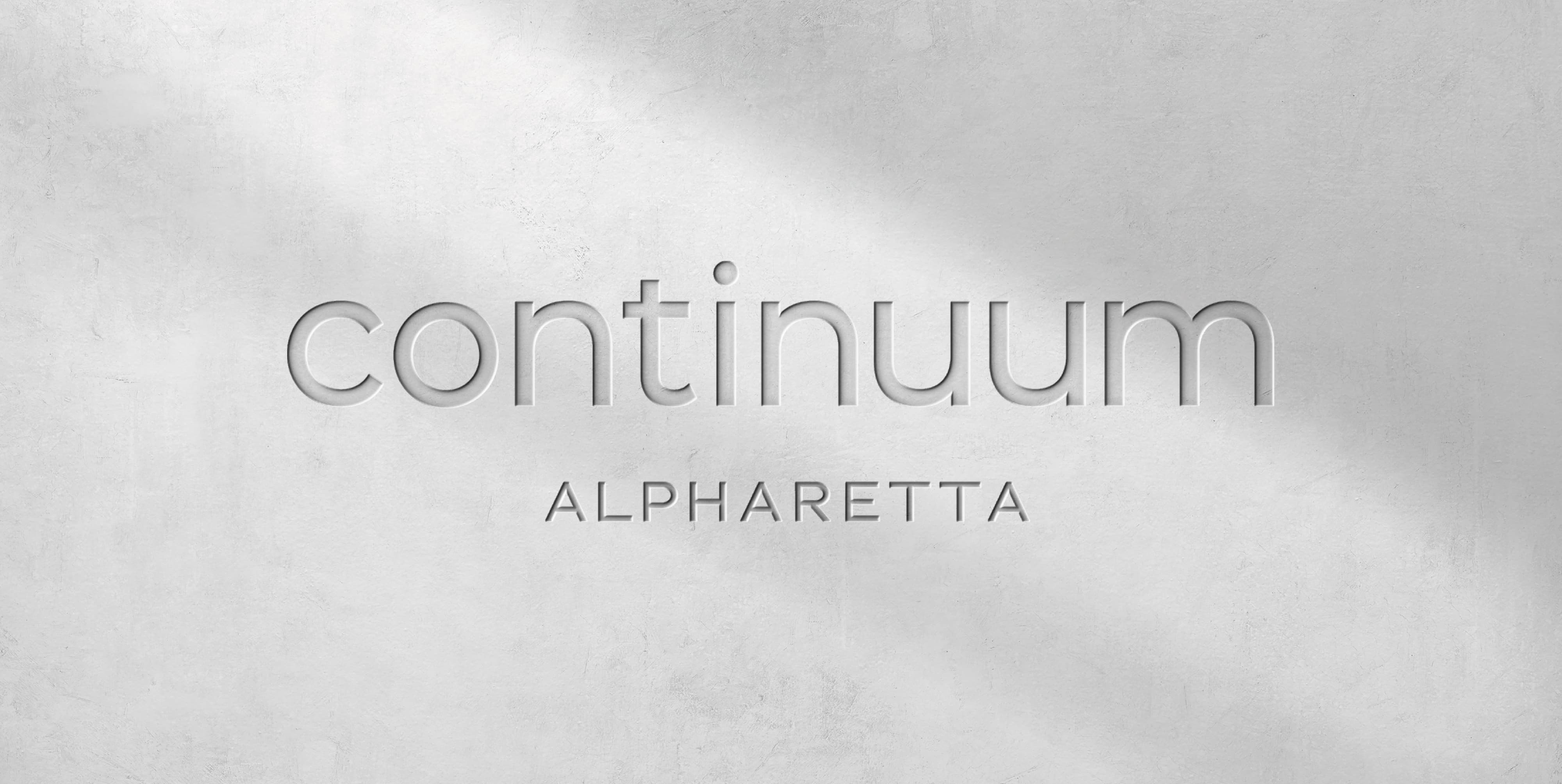 RSM Design visual identity and branding design for Continuum Alpharetta, featuring a close-up mockup of the Continuum Alpharetta logo with routed or debossed letterforms rendered on a soft concrete textured surface, showcasing the refined typographic character and material application of the brand identity system developed as part of a comprehensive environmental graphic design and branded environment program for a mixed-use workplace, health and wellness, and residential community development.
