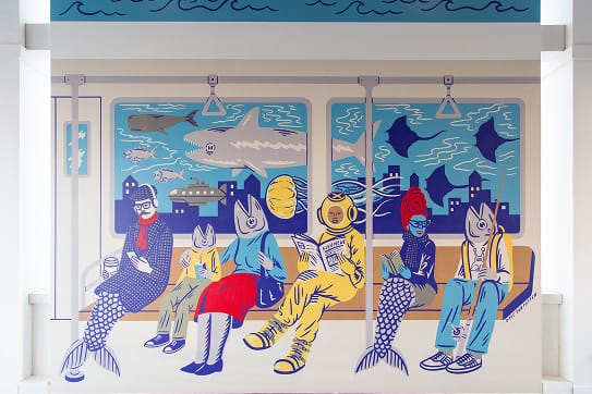 Large-scale interior mural at 2nd & PCH in Long Beach, California, depicting whimsical illustrated figures with fish heads and a deep-sea diver riding a subway car surrounded by an underwater ocean scene with sharks and rays — an example of RSM Design's art programs and public art, placemaking, and specialty graphic design that creates moments of delight within a coastal waterfront retail and mixed-use destination.