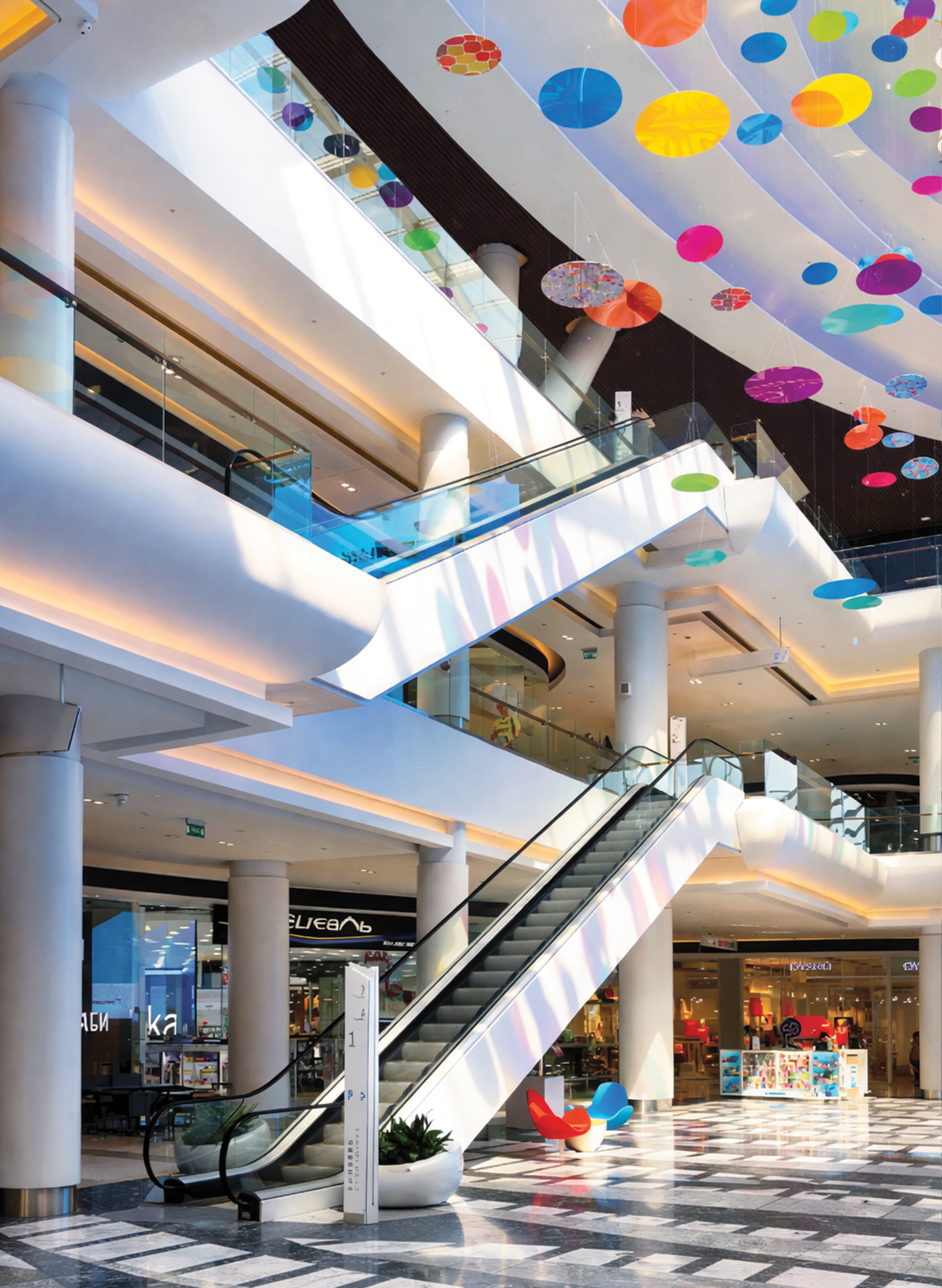 Multi-level international retail center interior atrium featuring a large-scale suspended colorful disc art installation, branded escalator experiential graphics, wayfinding kiosk, and patterned terrazzo flooring — RSM Design art programs, public art, experiential graphics, specialty graphics, wayfinding and signage design, and placemaking for retail, mixed-use, and international destinations.
