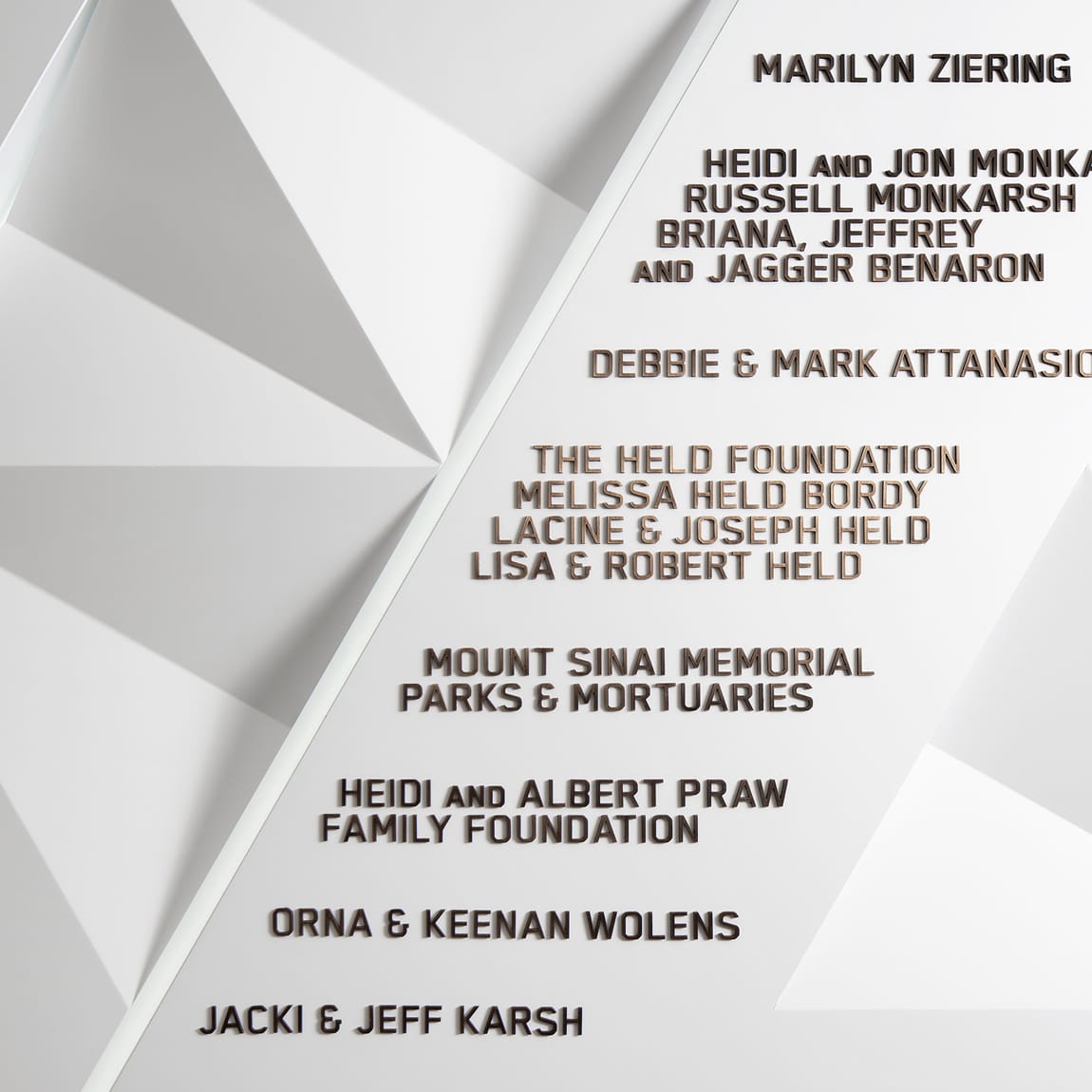 Detail view of an architectural donor wall featuring raised bronze three-dimensional lettering on white geometric faceted relief panels, recognizing contributors including Marilyn Ziering, Heidi and Jon Monkarsh, Russell Monkarsh, Debbie and Mark Attanasio, the Held Foundation, Melissa Held Bordy, Mount Sinai Memorial Parks and Mortuaries, Heidi and Albert Praw Family Foundation, Orna and Keenan Wolens, and Jacki and Jeff Karsh