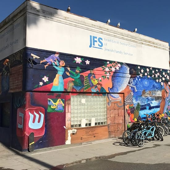Corner exterior of the JFS Israel Levin Senior Center of Jewish Family Service featuring a vibrant large-scale community mural depicting cultural and family scenes including a dreidel, figures dancing, and coastal imagery, with rental bicycles parked along the sidewalk
